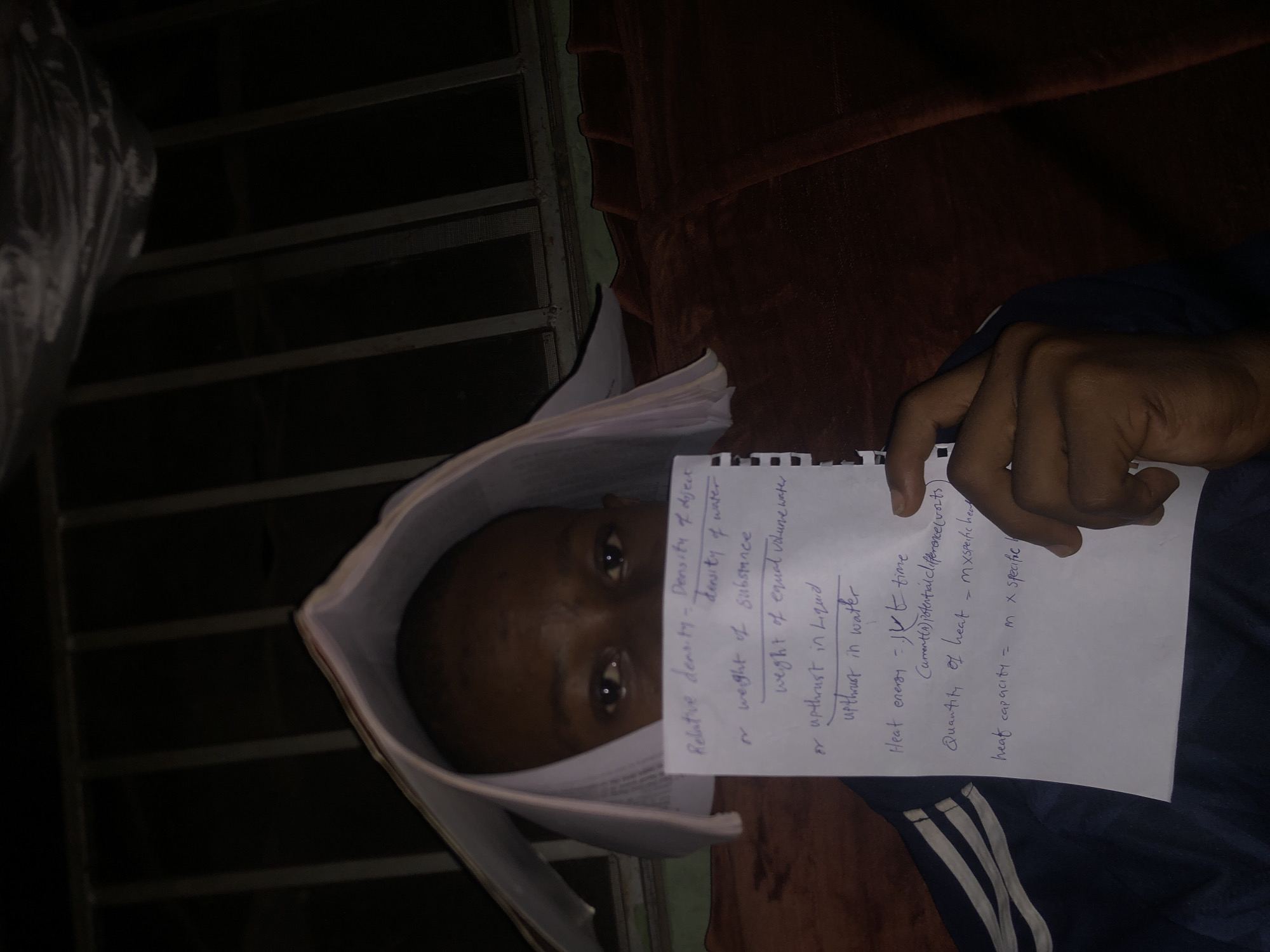 A person is lying on a surface that appears to be a bed or couch, holding a piece of paper with handwritten notes. The notes on the paper relate to physics concepts, including "Relative density", "Heat energy", "Quantity of heat", and "heat capacity." The person is positioned near a window with metal bars. The location is Atani, Nigeria.