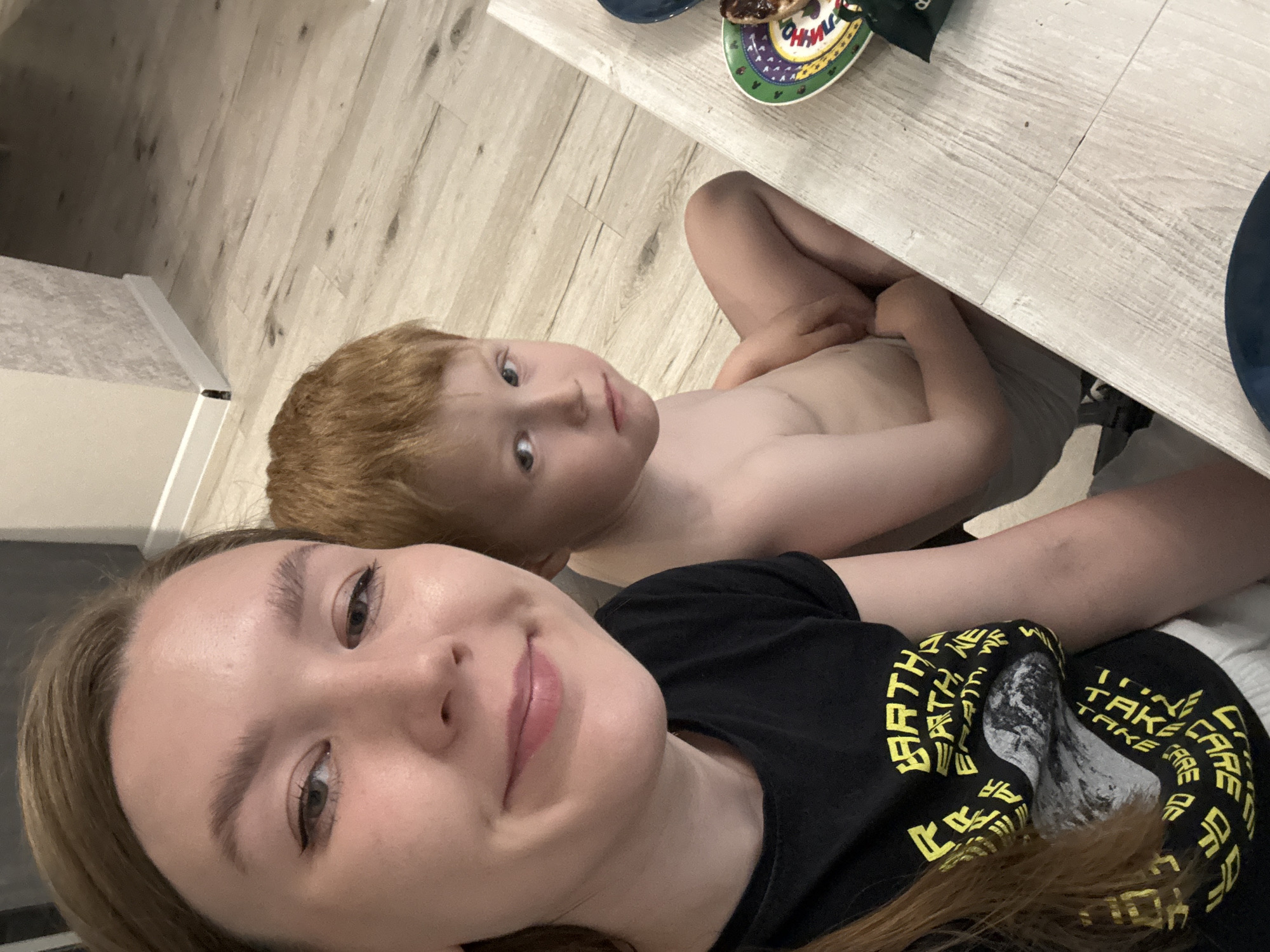 This casual, selfie-style photograph captures an adult woman and a young boy indoors, with a tilted perspective.
The woman, likely the photographer, occupies the foreground. She has light brown hair, fair skin, and a faint smile on her face. Her eyes are visible, enhanced with subtle eyeliner. She is wearing a black t-shirt adorned with yellow text. Clearly discernible words on her shirt include "EARTH" and "TAKE CARE," along with other fragmented text and a graphic of what appears to be a planet.
Behind the woman, and slightly above her, is a young boy. He has distinctive reddish-orange hair and light-colored eyes. He looks directly into the camera with a neutral, engaging expression. The boy is shirtless, wearing light grey shorts or underwear.
The setting appears to be a domestic indoor environment, possibly a kitchen or dining area. A light-colored wooden floor covers much of the background and parts of the foreground. A light-colored wooden table is visible in the upper right portion of the frame. On the table, there's a colorful plate with a decorative pattern and some Cyrillic text, which is indistinct but likely for a food item. A piece of bread or toast with a dark spread rests on this plate. A dark blue plate or bowl is partially visible next to it.
The image exudes a relaxed, intimate atmosphere between the two subjects. The lighting is consistent with indoor conditions, without strong indications of outdoor light or a specific time of day. The photograph was taken in Izhevsk, Russia.