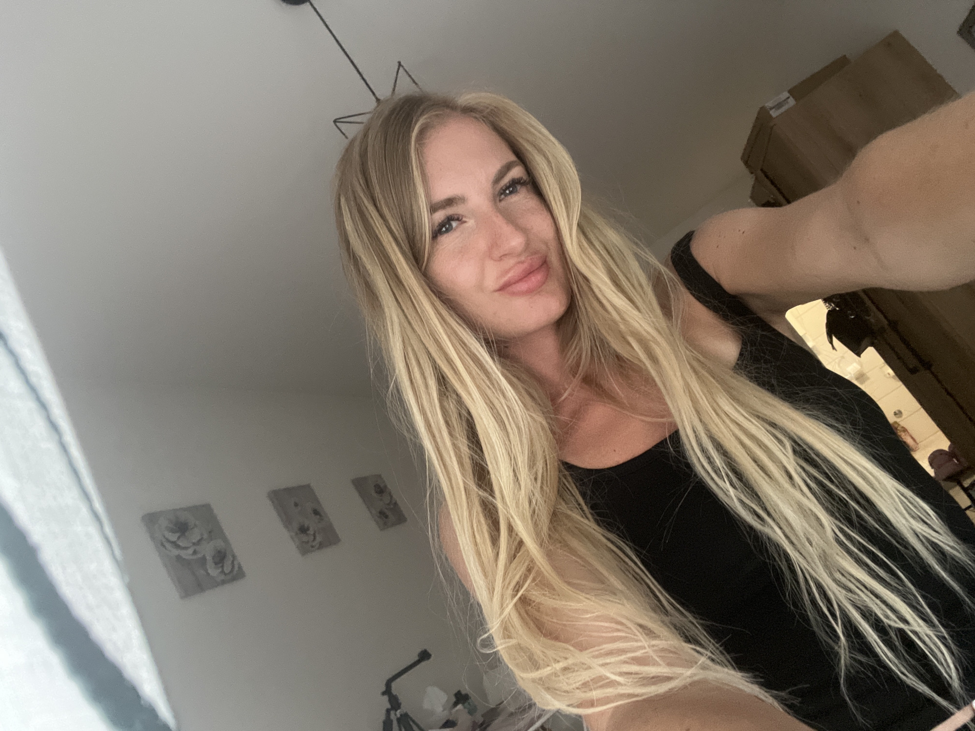 A medium shot captures a woman with long, blonde, wavy hair and blue eyes, looking directly at the camera. She is wearing a black tank top and her head is tilted to the right. The background is a plain, light-colored wall with three framed pictures of flowers mounted on it, suggesting an indoor setting. A portion of a light fixture is visible in the upper right corner. There is no indication of the city of Budva, Montenegro in the image.