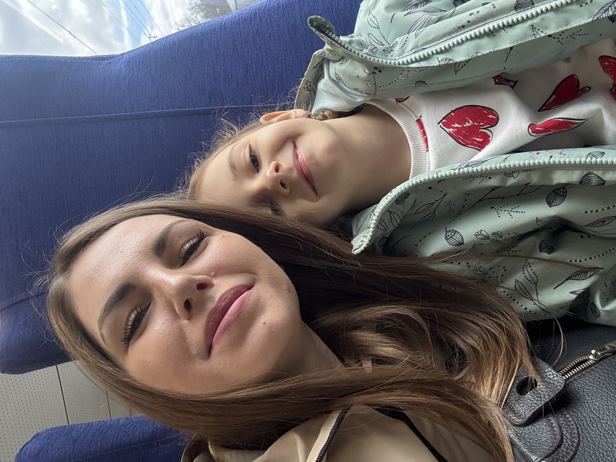 A selfie taken in what appears to be public transportation shows an adult female and a young girl. The adult female is lying on a blue seat, with her head close to the camera. She has long, brown hair and is wearing a beige jacket. The young girl is positioned slightly behind and above the adult female, also lying down. She is wearing a white sweater with red heart drawings and a patterned green jacket. The background includes a glimpse of the outside through a window. A black handbag is partially visible near the adult female. No evidence indicates the location is in Покровка, Russia.