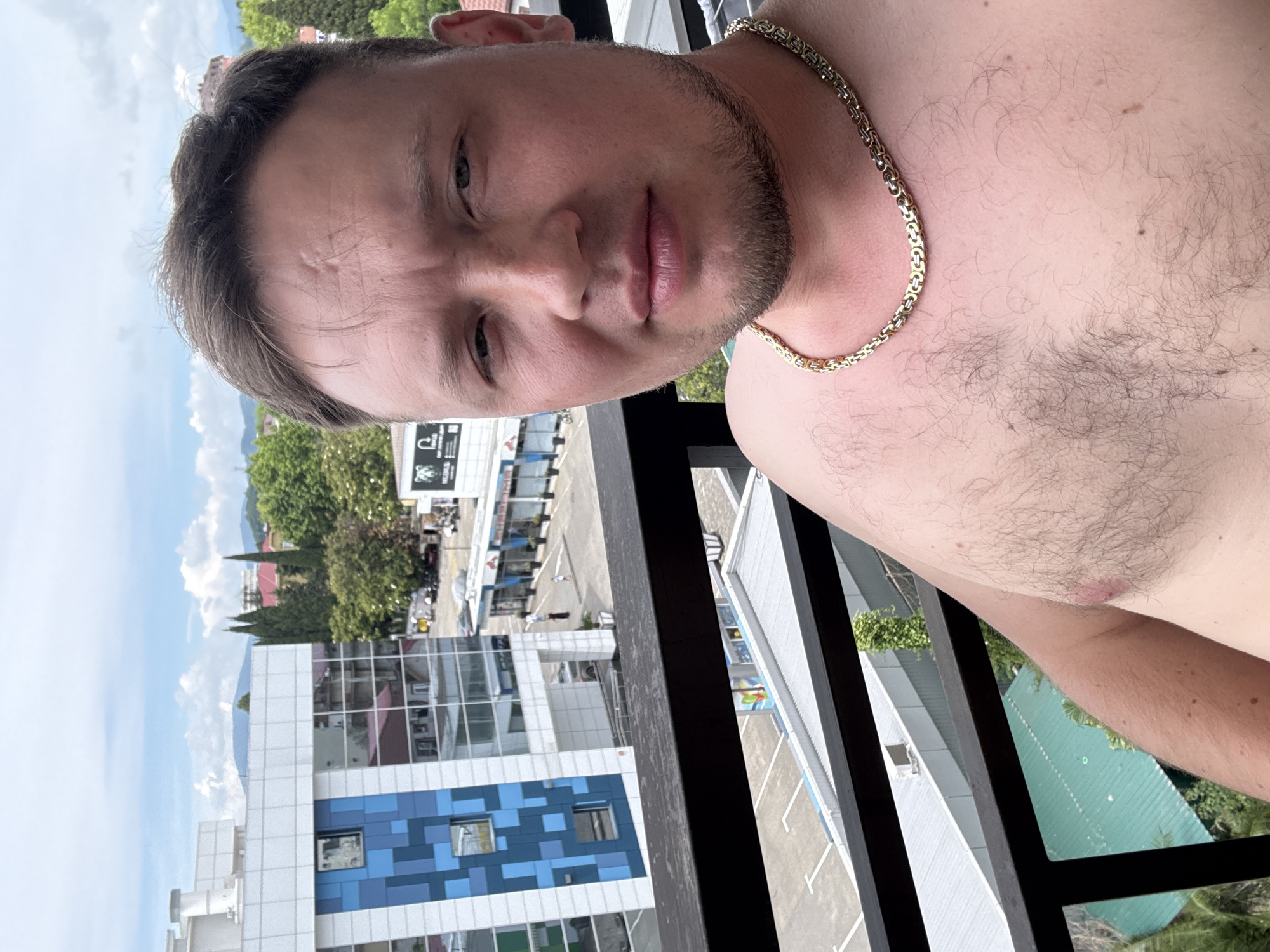 A man is taking a selfie on a balcony overlooking a city street in Sochi, Russia. He is wearing a gold chain around his neck and is shirtless. In the background, there are buildings, trees, and a cloudy sky.