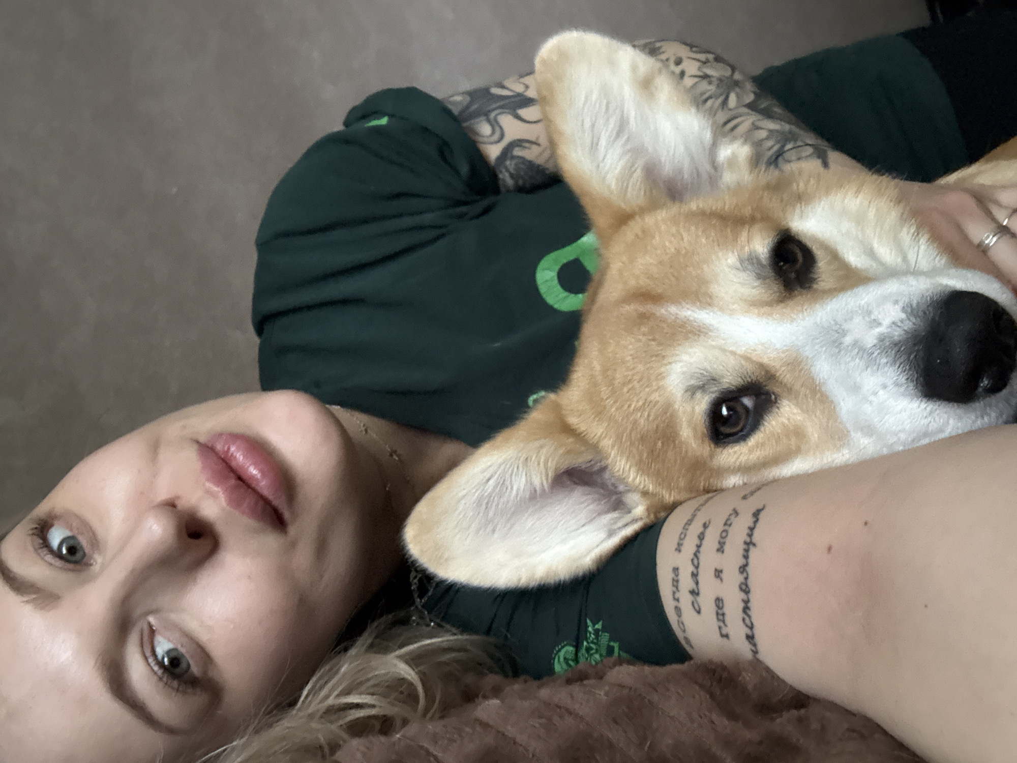 A woman with light skin and blonde hair lies on a brown surface, with a corgi dog nestled on her arm. The dog has a brown and white coat. The woman wears a dark green t-shirt with a green graphic. Tattoos are visible on her arm and upper chest. The scene's location in Borzya, Russia, cannot be determined based on the visual information.
