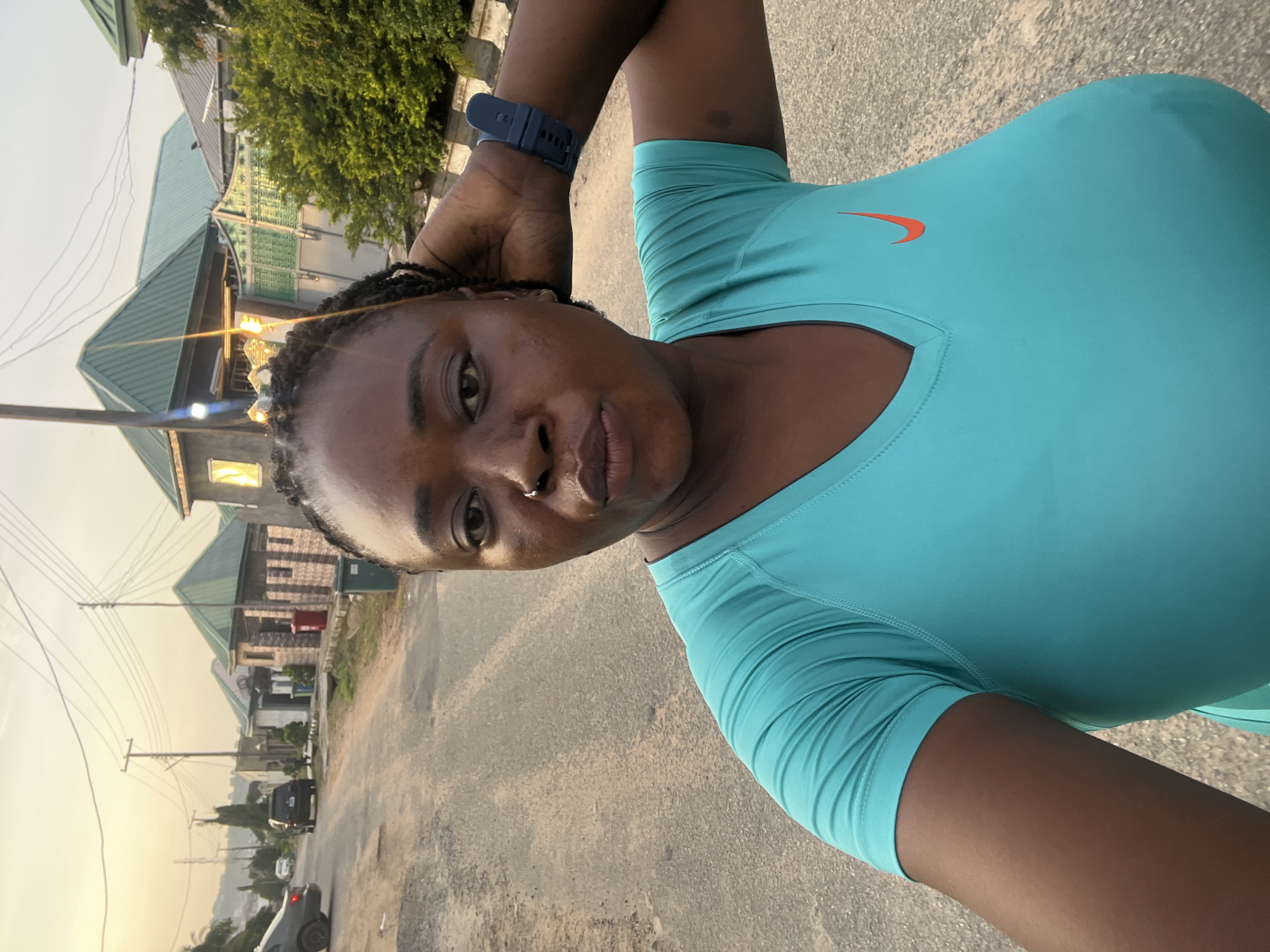 A medium shot captures a Black woman with braided hair and a nose ring from the chest up, facing the camera with her left hand behind her head. She is wearing a teal t-shirt with an orange Nike logo on the left side of her chest and a dark blue smartwatch on her left wrist. The woman is standing outdoors on a paved road with houses and trees in the background, suggesting a residential area. The sky is overcast with a warm glow, indicative of either sunrise or sunset. The scene appears to be in Abuja, Nigeria, given the architectural style of the houses and the general environment.