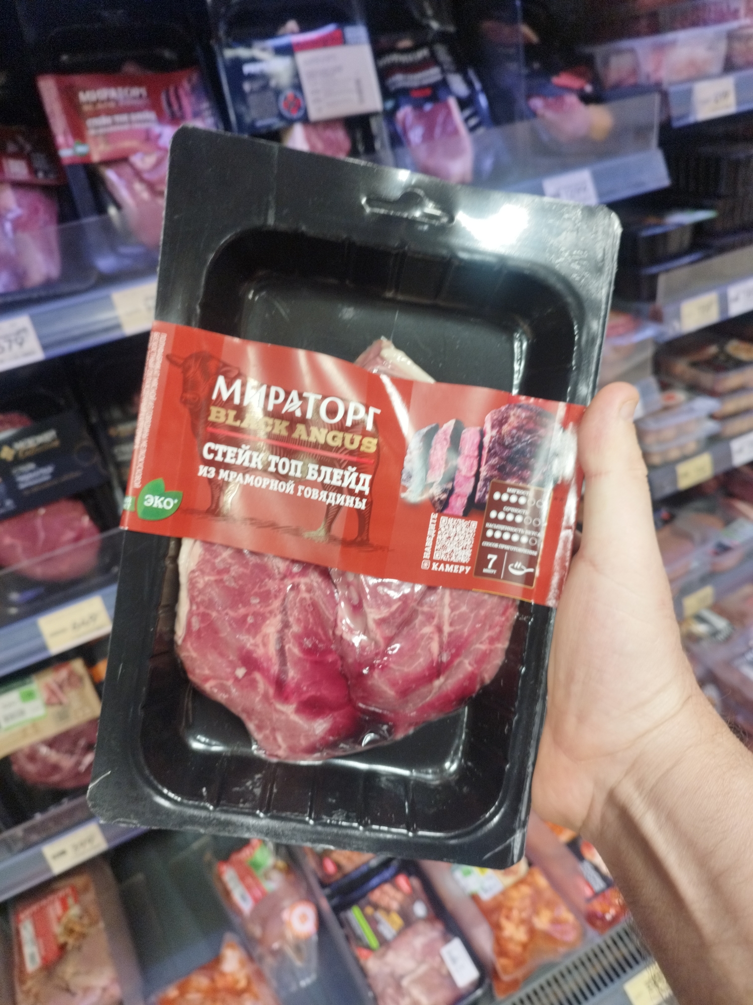 In a retail environment, a hand holds a black plastic container containing raw beef steaks, identified by a red label as "МИРАТОРГ BLACK ANGUS СТЕЙК ТОП БЛЕЙД ИЗ МРАМОРНОЙ ГОВЯДИНЫ" (Miratorg Black Angus Top Blade Steak from Marbled Beef). The label also features an image of a bull, a stylized depiction of sliced steak, and a QR code. The background displays shelves stocked with other packaged meat products. The overall scene suggests a typical supermarket aisle. This image was captured in Balashikha, Russia.