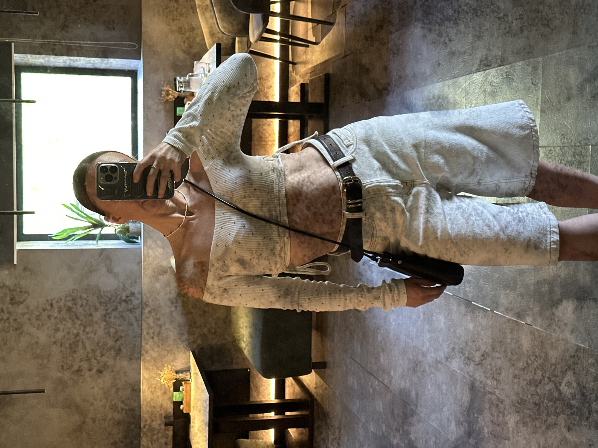 A person is taking a selfie in an indoor environment, possibly a café or restaurant. The person is wearing a light-colored, long-sleeved top, light-colored shorts with a dark belt, and a crossbody bag. They are holding a smartphone with a dark-colored case. The background includes chairs, tables, a window, and decorative items. The location information is not available in the image.