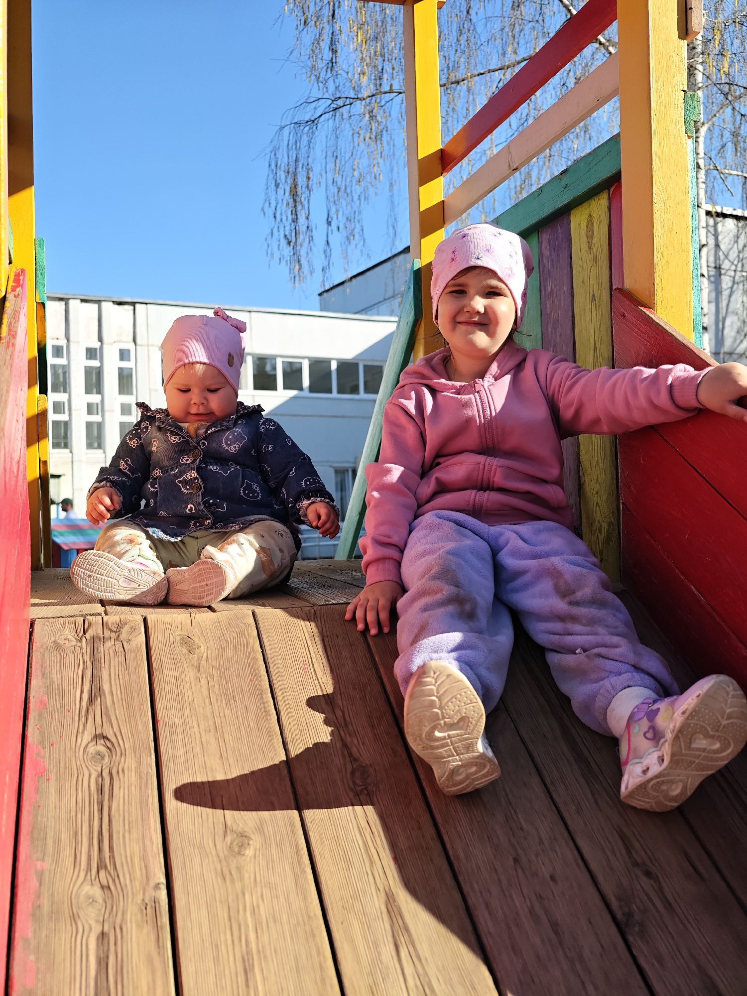 Two young girls, both wearing pink hats, are sitting on a wooden playground structure. The older girl is wearing a pink jacket and lavender pants and sits toward the right, while the younger girl, dressed in a dark jacket with light pants, sits on the left. The playground equipment is painted in bright colors, including yellow, red, green, and blue. In the background, a white building is visible with a blue sky above. The location is Chaikovsky, Russia.