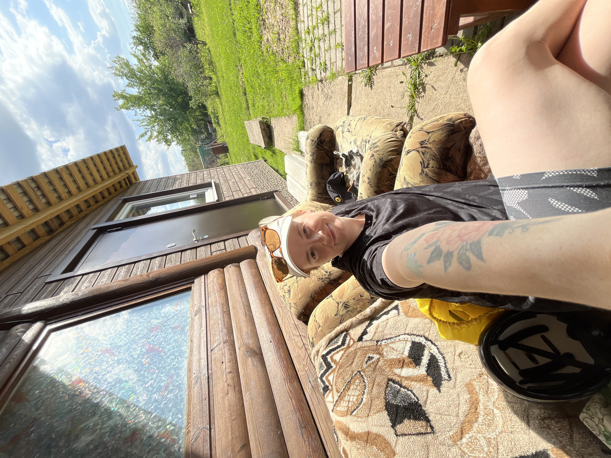 A person is sitting outdoors on a patterned couch next to a wooden building. The building has a window with a textured film applied to it and a brown door. The person is wearing a black shirt, patterned shorts, a white headband, and sunglasses. There is a black object on the couch next to the person. In the background, there is green grass and trees under a partly cloudy sky. The location is Chaikovsky, Russia.