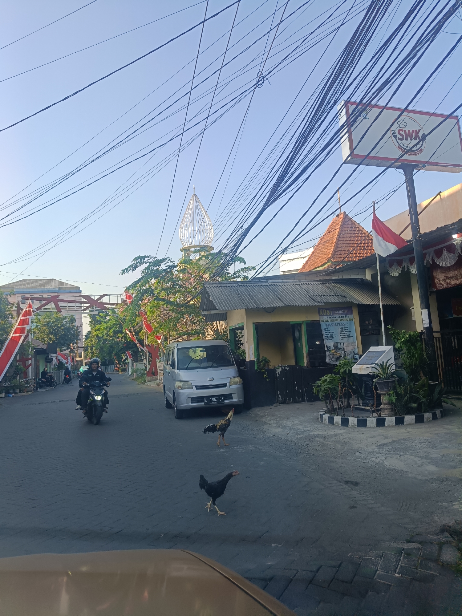 The image depicts a street scene in Surabaya, Indonesia, with a mix of residential and commercial buildings. Overhead, a dense network of electrical wires crisscrosses the frame against a clear blue sky. In the foreground, a portion of a car's hood is visible. The street itself is paved with interlocking bricks. Two chickens are present on the road, one black and one speckled.
To the right, a light-colored van with license plate "L 1384 CAI" is parked. Adjacent to the van is a building with a reddish-brown tiled roof and a sign that reads "KOST EXCLUSIVE" and lists amenities like "AC/TV", "Kamar Mandi Dalam" (en-suite bathroom), "Kasur Busa" (foam mattress), and "Meja Belajar" (study desk). A small monument or plaque is situated in front of this building, surrounded by potted plants. Further to the right, a sign for "SWK" (Siwak Akeglu) is visible, featuring a chef's hat and knife logo. An Indonesian flag, red and white, waves from a pole in front of a building.
On the left side of the street, a motorcyclist wearing a helmet is riding towards the viewer. Several other motorcycles are parked along the sidewalk. Red and white Indonesian flags and pennants are displayed on poles along the street, suggesting a national celebration or event. In the background, a tall, modern building with a distinctive white, cage-like dome structure is visible, possibly a landmark or religious building. Green trees are scattered throughout the scene. The lighting suggests it is late afternoon or early morning, with a soft, natural light casting shadows.