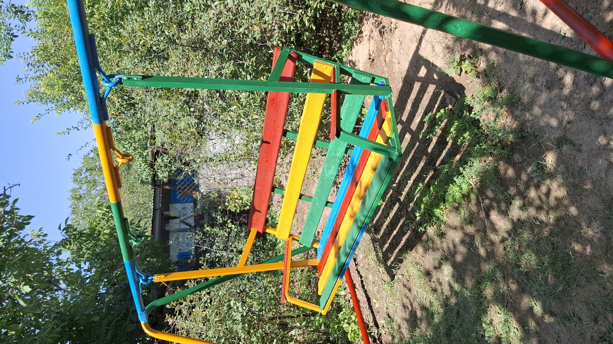 A brightly colored swing set with a multi-colored seat is positioned in a yard, likely in the city of Шұбарсу, Kazakhstan. The swing set features a blue and yellow horizontal bar, supported by green vertical poles. The seat itself is made of wooden planks painted in alternating colors of red, yellow, green, and blue. The background consists of dense green foliage and trees, with a partially visible building structure behind the swing. The ground beneath the swing is a mixture of dirt and grass, with dappled sunlight casting shadows from the swing set onto the ground. No people are present in the image.