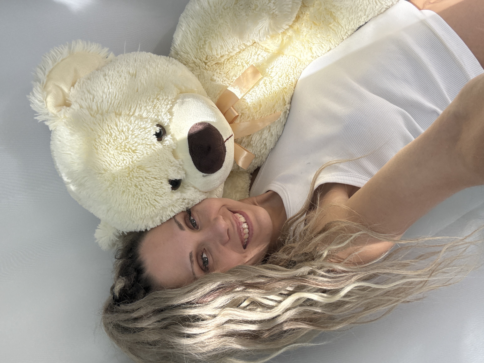 A blonde woman with light skin and long, wavy blonde hair is lying on her side, embracing a large, cream-colored teddy bear. She is looking at the camera with a smile, her head resting on the teddy bear's shoulder. She is wearing a white ribbed tank top. The teddy bear has a brown nose and a light peach ribbon tied around its neck. The background is a plain white surface, likely a wall or a bed. There is bright lighting casting shadows. The image appears to be a selfie taken indoors. There is no indication of the city of Zhukovo, Russia in the image.