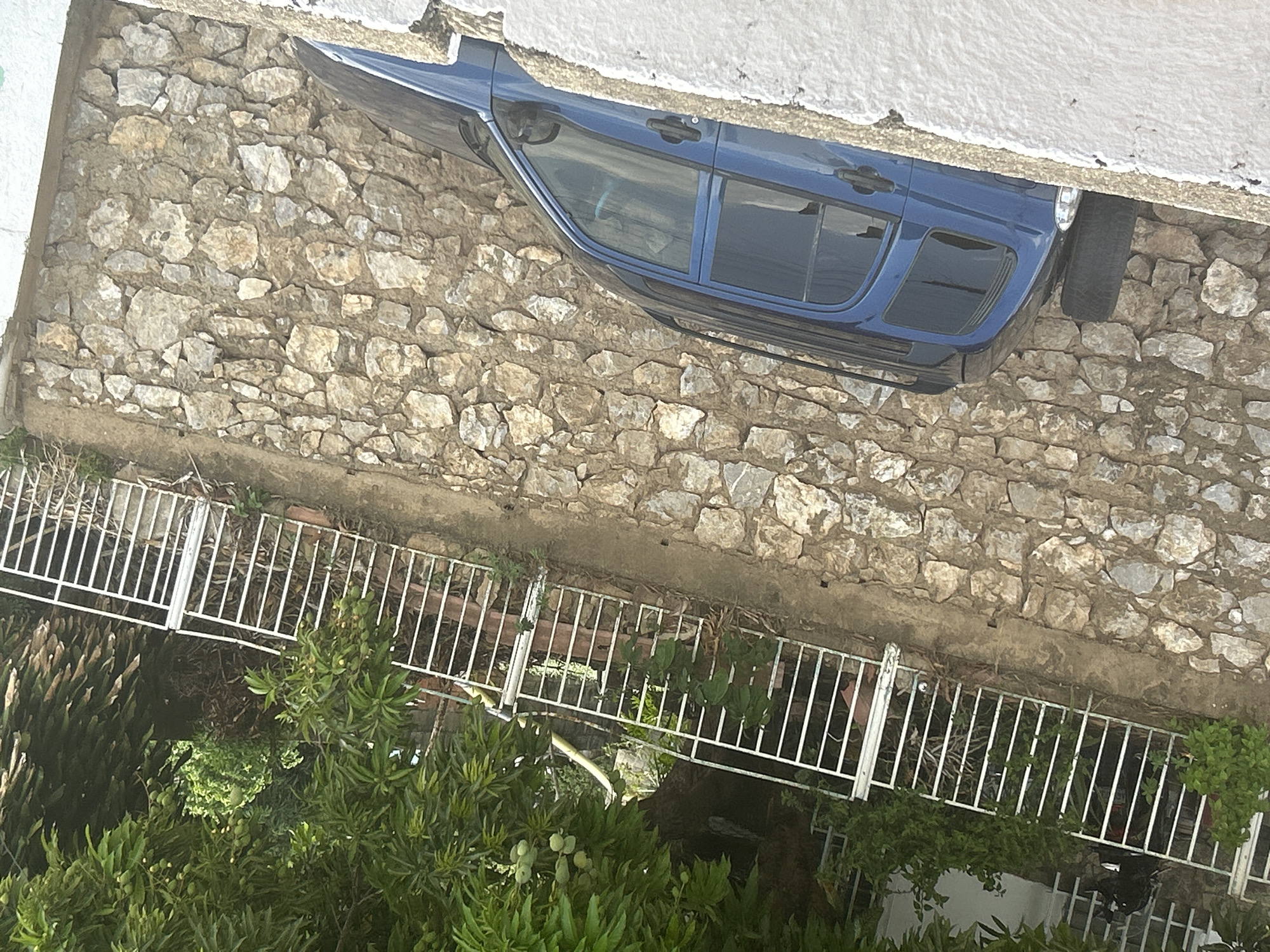 The image depicts an elevated perspective of a scene in San Juan de Los Morros, Venezuela. A blue vehicle is parked adjacent to a stone wall that runs along a property line defined by a white metal fence. Vegetation, including trees and shrubs, grows below the fence line. A white building structure is partially visible above the vehicle and wall.