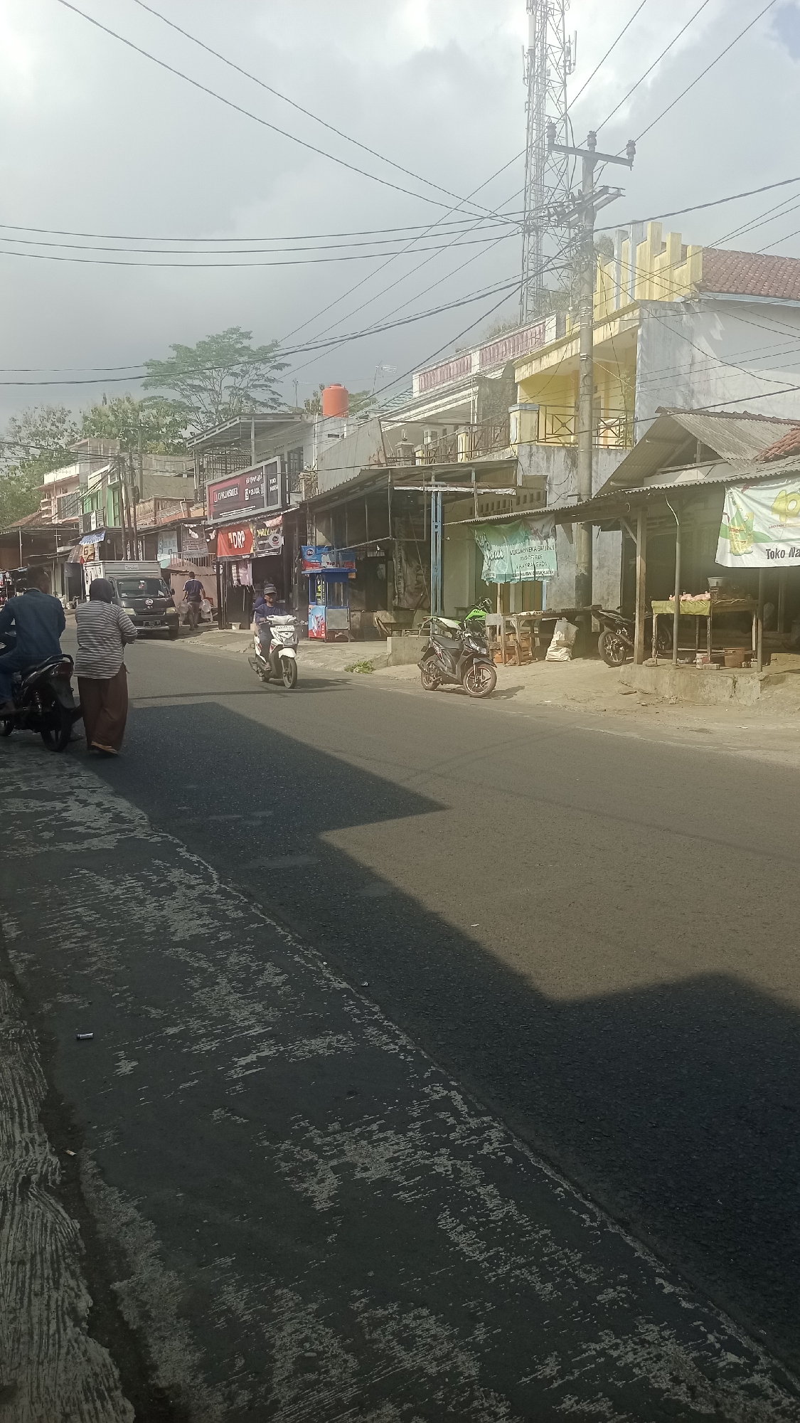 This image depicts a street scene in Karyamandala, Indonesia, on a slightly overcast day. The road is paved and shows signs of wear, with patches of lighter material and debris. On the right side of the road, there are several small shops with covered awnings and open fronts, displaying various goods. One shop has a green banner with text. A few motorcycles are parked or in motion on the street. On the left side of the road, there are more buildings and shops. A few people are visible, including individuals on motorcycles and walking. A dark-colored van is parked in the distance. Power lines crisscross above the scene, and a cell tower is prominent in the background. The overall impression is of a typical commercial street in a smaller Indonesian town.