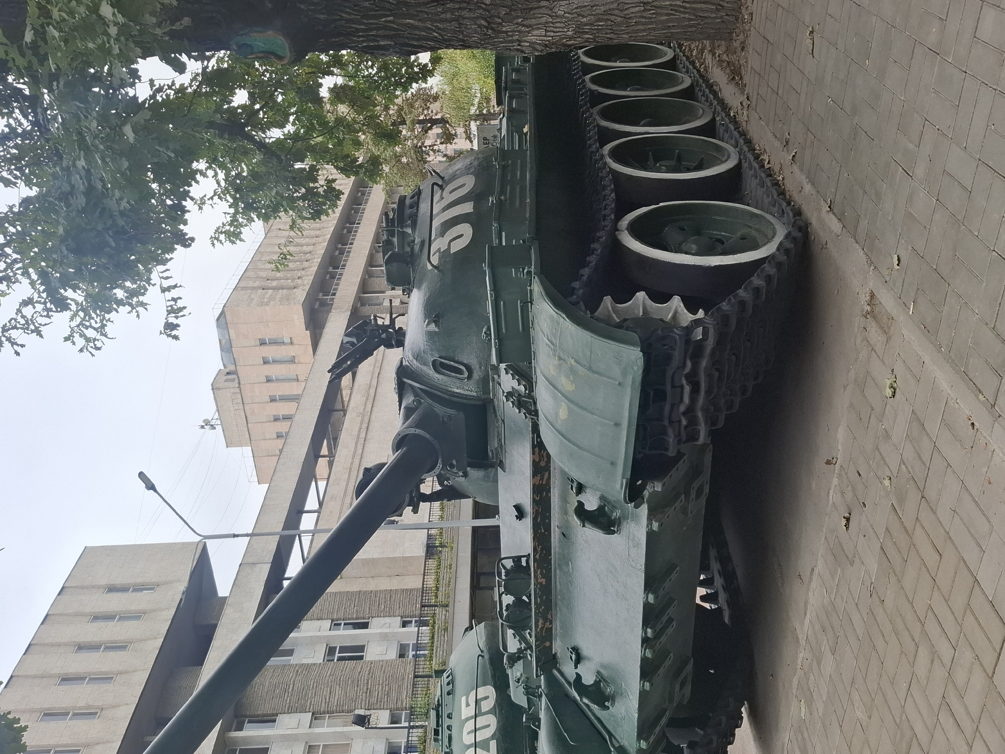 The image shows two green tanks parked on a brick sidewalk in front of a Brutalist-style building. The tank in the foreground has the number "376" painted on its turret. The tank in the background has the number "205" painted on its side. A large tree with green leaves is on the right side of the image. The sky is overcast. The building appears to be a government or educational institution. This scene is consistent with the Memorial to the Defenders of the Fatherland in the city of Almaty, Kazakhstan, which features military equipment on display.