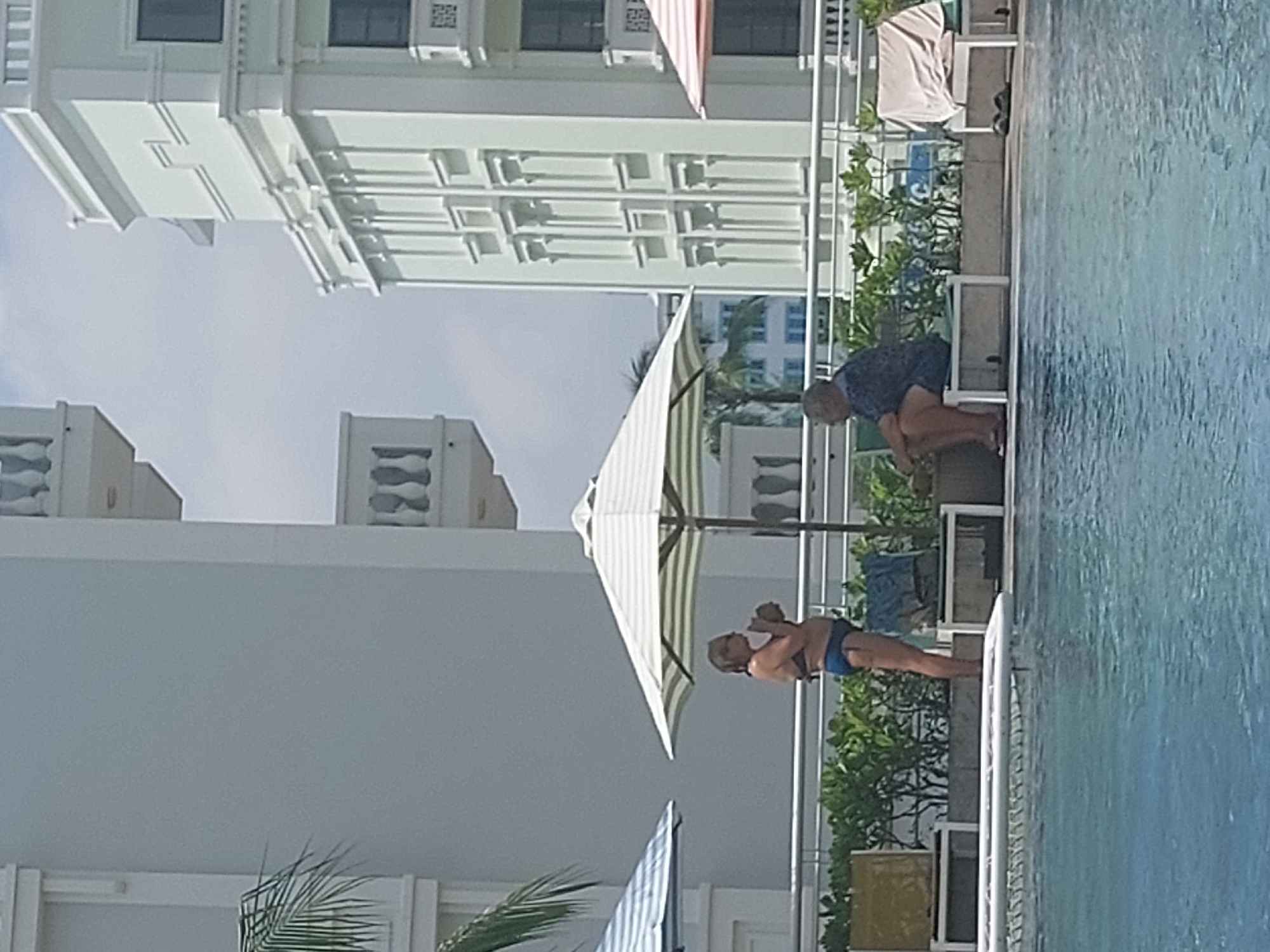A woman in a blue bikini stands under a striped umbrella by a swimming pool. She is holding a drink. To her right, a man in a dark shirt and shorts sits on a bench, facing away from the camera. Behind them, lush green foliage lines the edge of the pool area, with a white railing separating it from the pool water. In the background, modern buildings with decorative white facades and balconies are visible against a hazy sky. The scene appears to be a resort or hotel in Phú Quốc, Vietnam, given the tropical vegetation and architectural style.