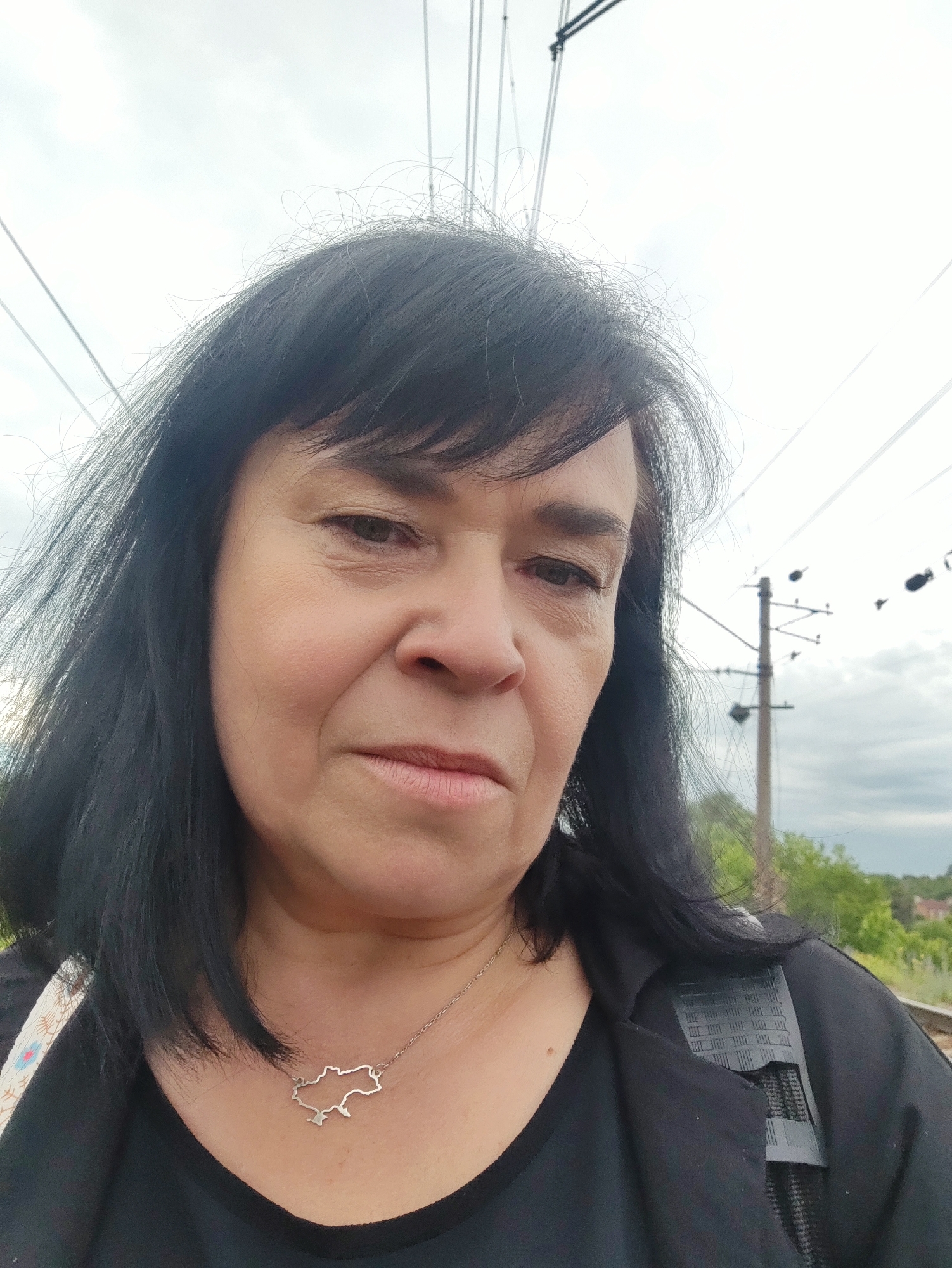 A woman with dark hair and a black top is posing for a selfie outdoors. She is wearing a silver necklace with a pendant shaped like the outline of Ukraine. In the background, there are power lines and a utility pole, suggesting the presence of a railway or power infrastructure. The sky is overcast, and there are hints of greenery in the distance.