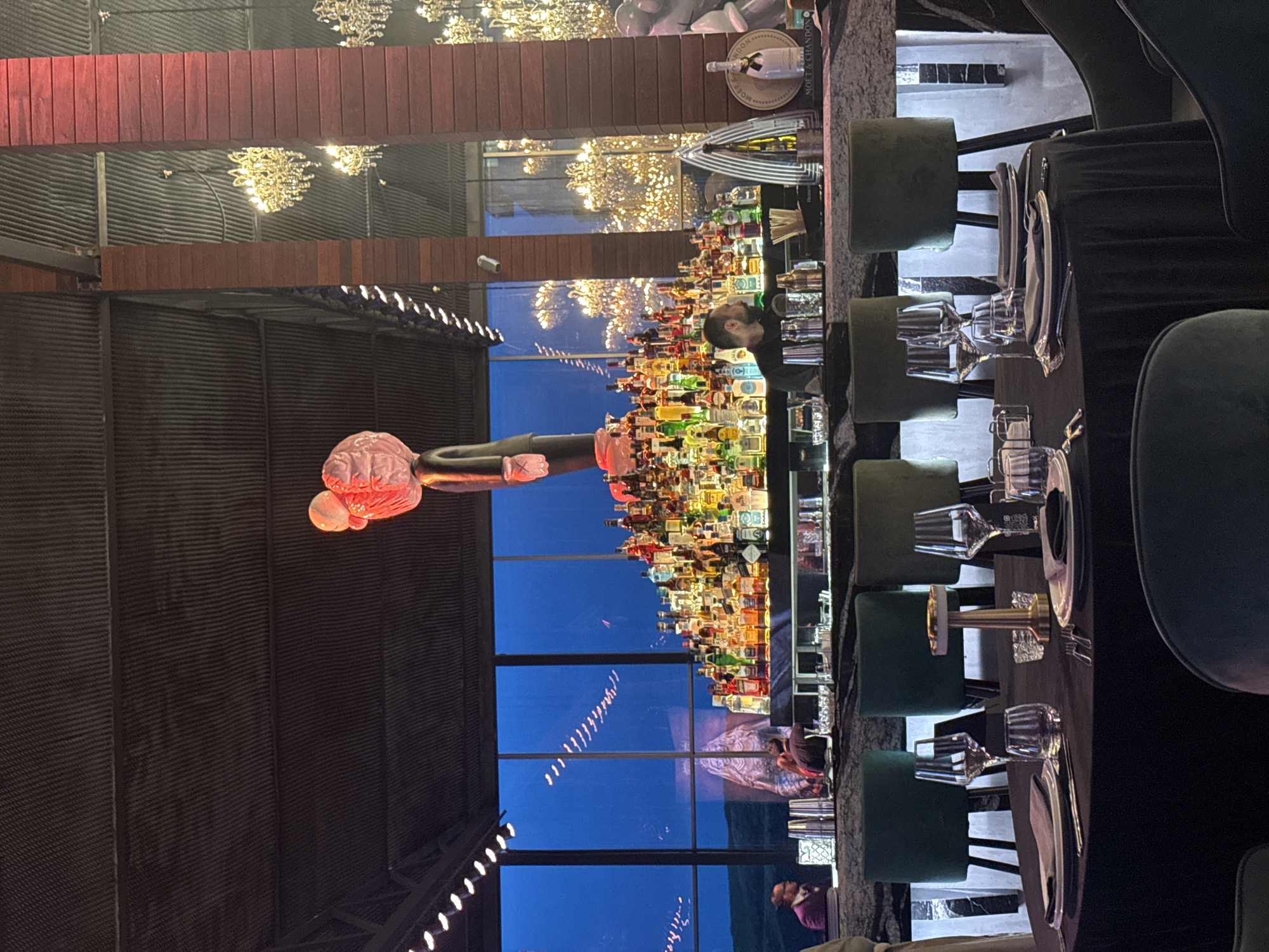 The image shows the interior of a restaurant in Yerevan, Armenia. A long bar is lined with numerous bottles of alcohol. A bartender is visible behind the bar. The dining area features tables set with plates, silverware, and glassware. Chairs are placed around the tables. Outside the restaurant, a pink sculpture is visible through large windows. The building has an industrial design, with exposed metal beams. Chandeliers hang from the ceiling.