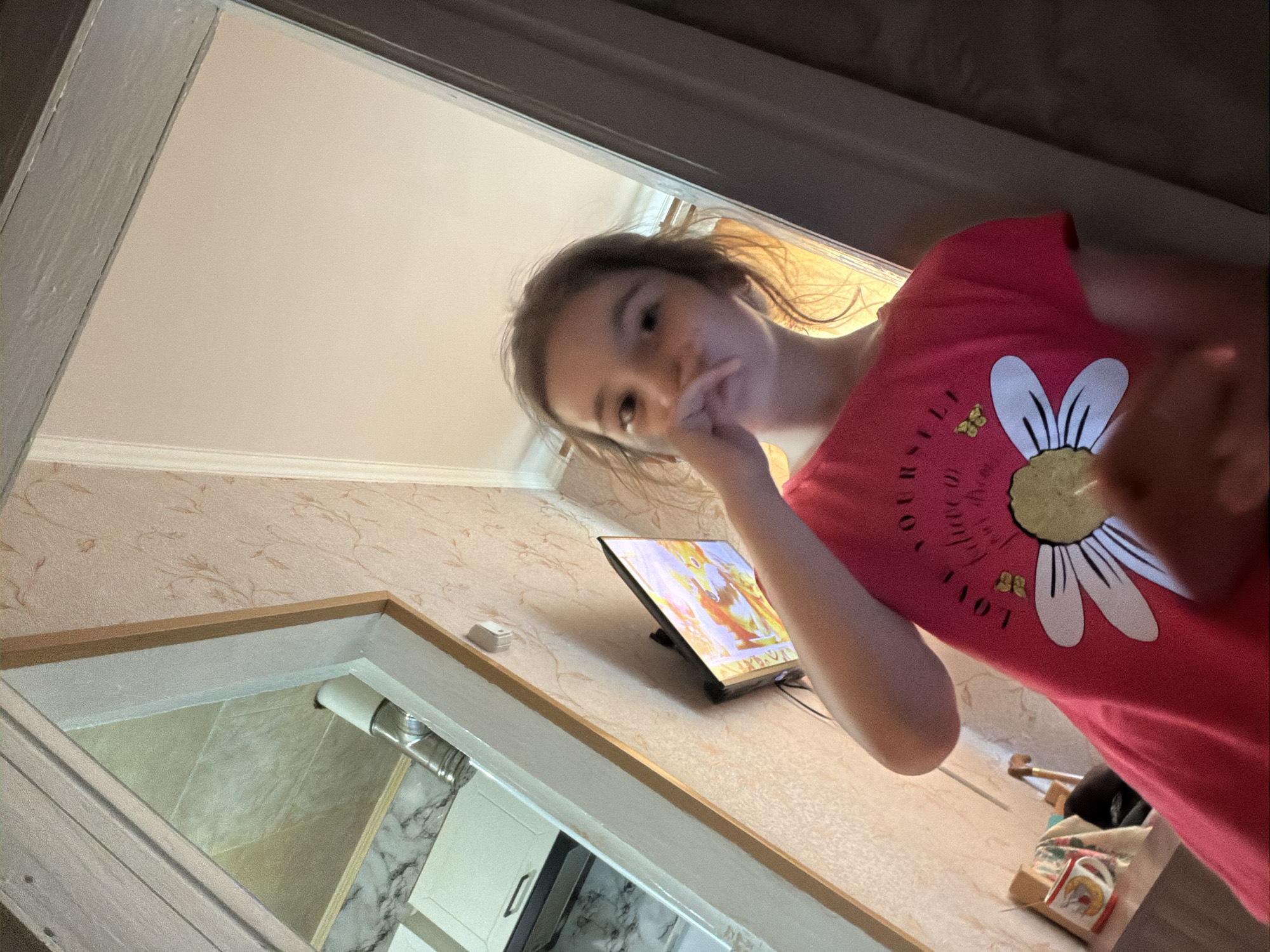 The image shows a young girl in a pink t-shirt with a daisy design and the text "love yourself believe in" printed on it. She is standing in what appears to be a doorway or hallway, with a mirror reflecting part of a kitchen area on the left. A television is mounted on the wall to the right of the mirror, displaying a colorful image. The girl's hand is raised to her face, obscuring her mouth. The lighting suggests an indoor environment, possibly during the daytime. The location is identified as Tambov, Russia, however, there are no specific visual cues in the image to confirm this location.
