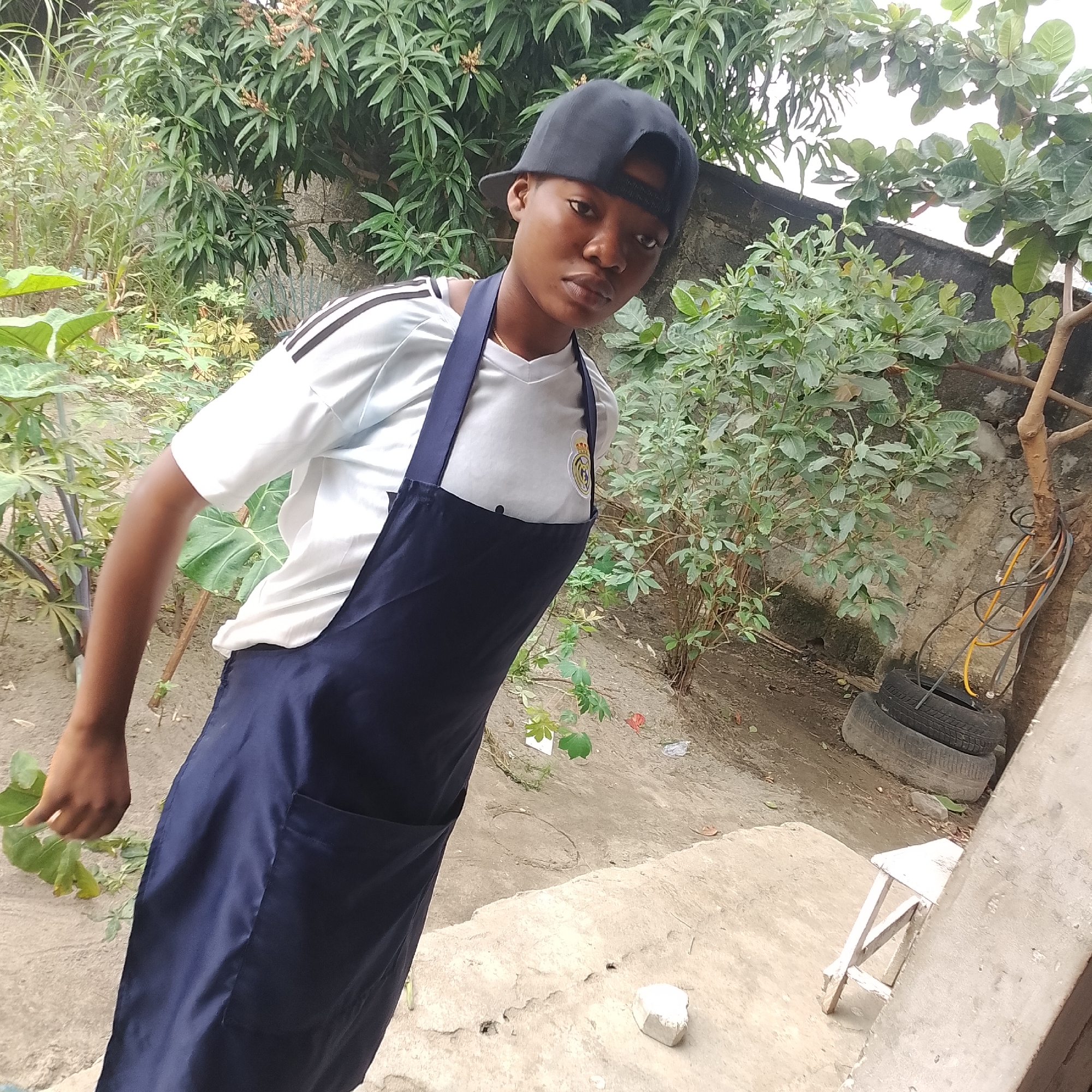 A young person wearing a dark blue apron over a white t-shirt with black stripes on the sleeves and a black baseball cap is standing outdoors in what appears to be a residential backyard in Ado, Nigeria. The person is looking towards the camera. The environment is characterized by lush vegetation, including trees and smaller plants. In the background, there is a concrete wall and some stacked tires. A small wooden stool is visible to the right of the image. The ground is dirt and there are some scattered leaves and debris. The lighting suggests it is daytime.