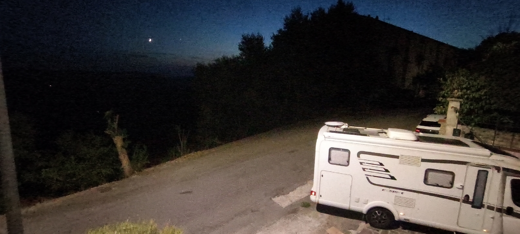 The image depicts a nighttime scene in Campagnatico, Italy. A white recreational vehicle (RV) is parked on a paved surface that appears to be a road or driveway. The RV is positioned on the right side of the frame, facing left and slightly towards the viewer. The vehicle has several windows, vents, and graphics on its side, including the model designation "E 515-t". A small satellite dish is visible on the roof of the RV.
Behind the RV, the terrain slopes upward and is covered in dark foliage, indicative of a wooded area or hillside. A building or structure, possibly a stone wall, is partially visible on the far right. In the distance, a faint crescent moon and a bright star are visible in the deep blue twilight sky. The overall lighting suggests it is early evening or late night. The ground in the foreground is paved, with some grass or vegetation visible along the edges. No people are present in the image.