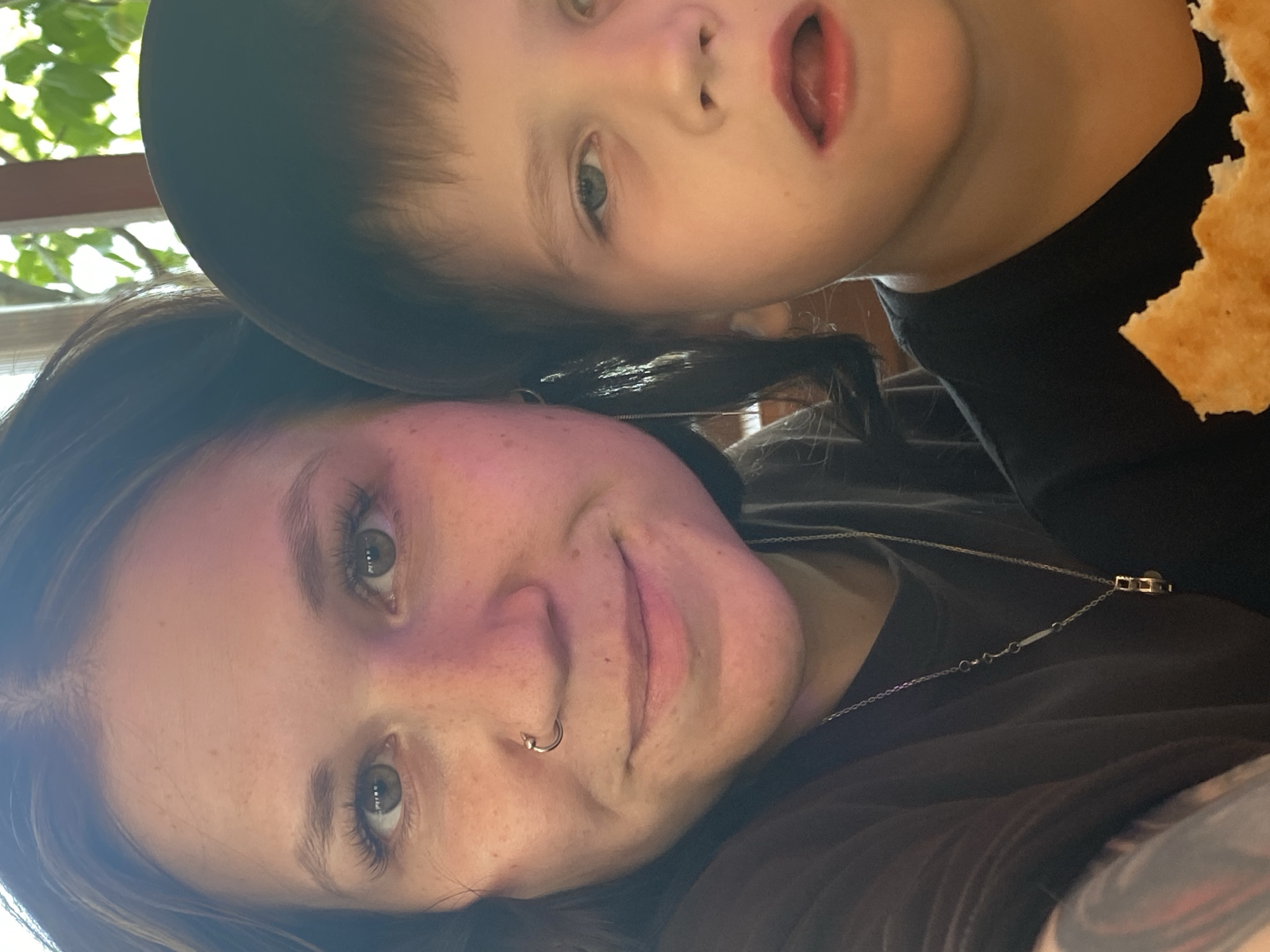 A selfie shows a woman and a young boy close together. The woman, in the foreground, has fair skin with freckles, green eyes, and a nose ring. She is wearing a dark-colored top and a silver necklace. Part of a tattoo is visible on her arm. The boy, in the background and slightly out of focus, has fair skin, light eyes, and dark hair. He is holding a piece of bread or a cracker. The background is blurred, with hints of greenery suggesting an outdoor setting. The photo may have been taken at the city of Chaikovsky, Russia.