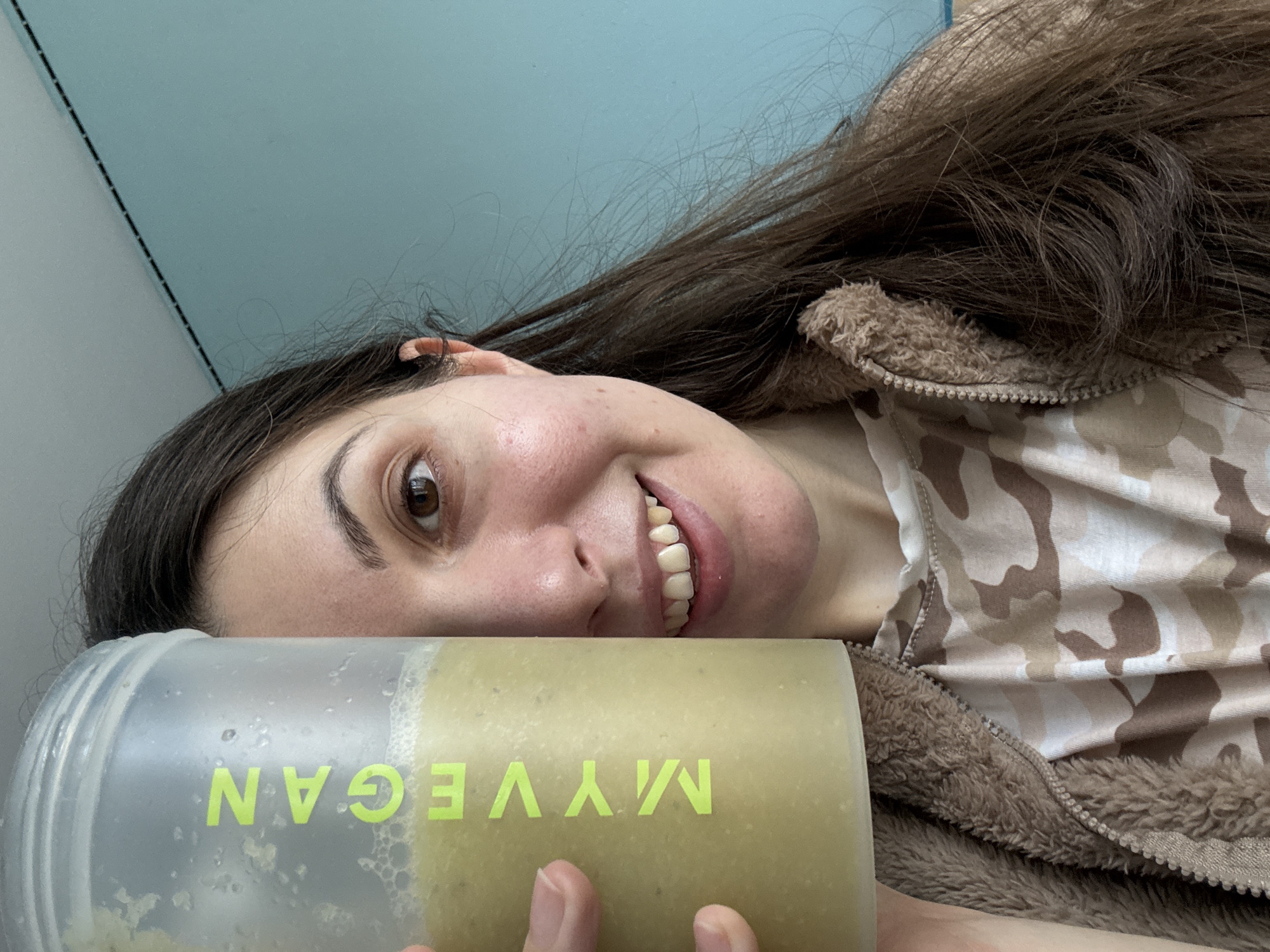 A woman is holding up a Myvegan protein shake. She is wearing a brown hooded sweatshirt with a camouflage pattern. The background is a light blue wall. The location is unknown.