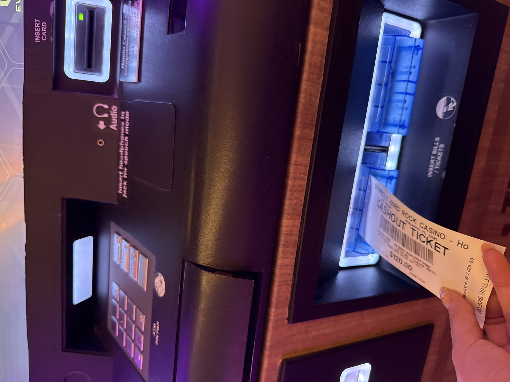 The image shows a close-up of a slot machine at the Seminole Hard Rock Casino in Hollywood, Florida. A hand is pulling a cashout ticket from the machine. The ticket indicates a payout of $120.00. The machine features slots for inserting a card and bills/tickets, as well as an ATM pin pad.