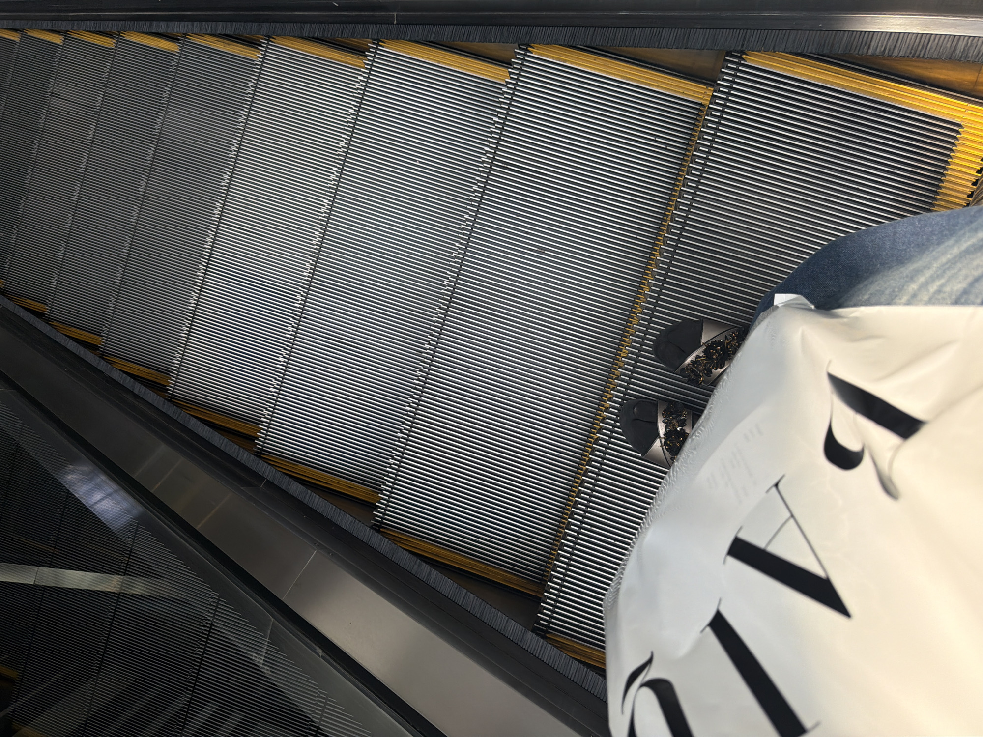 The image shows a first-person perspective of someone standing on an escalator. The escalator has metal steps with yellow accents along the edges, and a dark gray railing. In the foreground, a white shopping bag with large black lettering is partially visible. The person is wearing blue jeans and sandals adorned with sequins and floral embellishments. The scene appears to be in a modern indoor setting, potentially a shopping mall or commercial building in East Jakarta, Indonesia, given the context provided. The lighting suggests an indoor environment with artificial illumination.