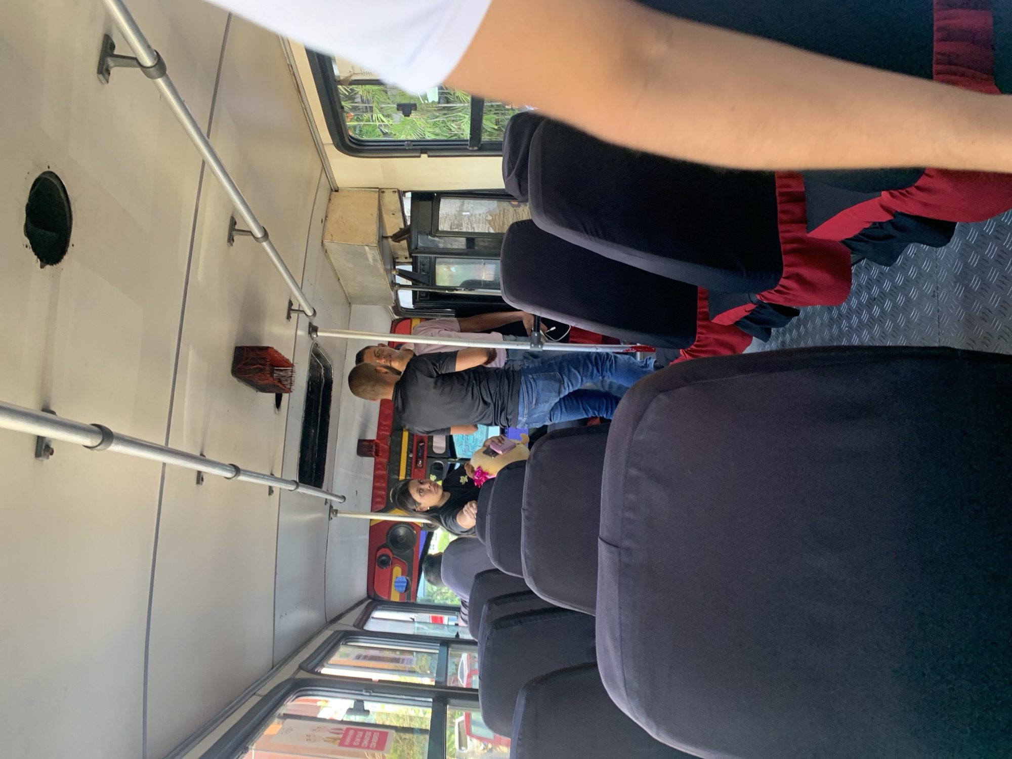 The image shows the interior of a public transport vehicle, possibly a bus or van, likely in Porlamar, Venezuela based on the presence of people and the general appearance of the interior. Rows of dark-colored, likely vinyl, seats occupy the majority of the frame, leading towards the front of the vehicle. At least two passengers are visible towards the front: a man wearing a dark shirt and jeans appears to be standing or walking in the aisle; and a woman with dark hair is seated. Other potential passengers are visible further down the aisle. The vehicle interior has light-colored walls, a metal handrail running along the side, and windows showing a glimpse of greenery outside. Some red material is seen on the seats closer to the camera.
