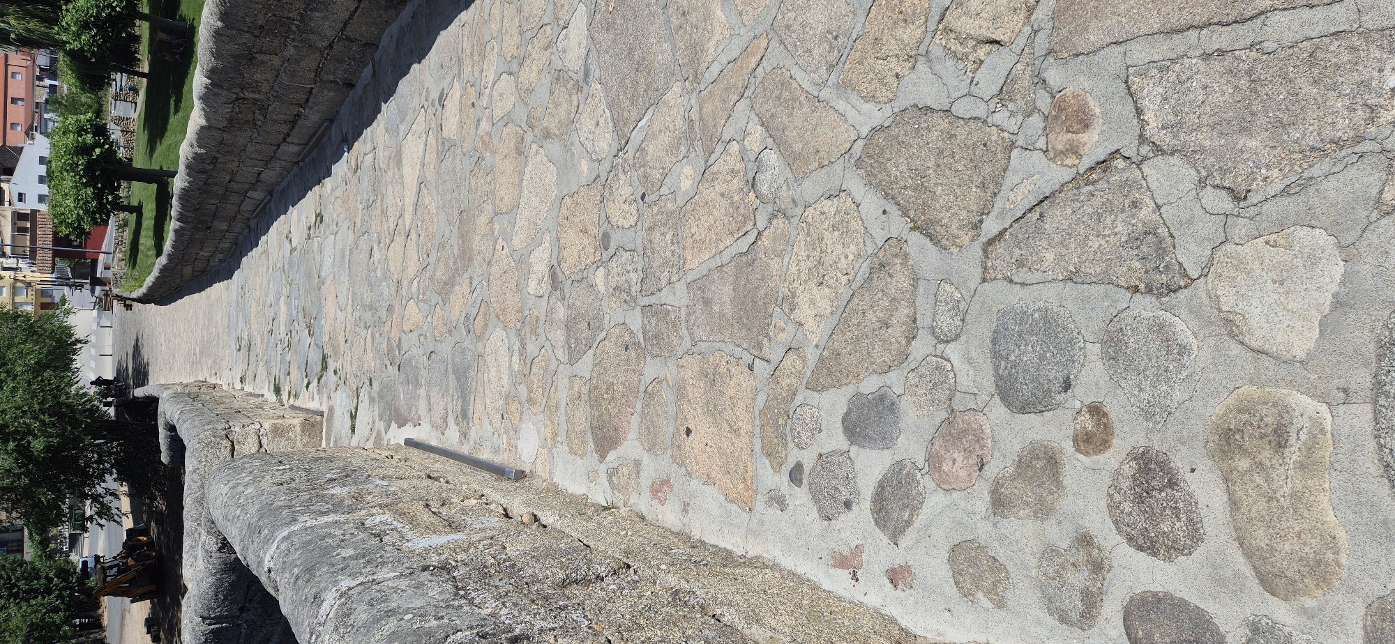 This is a high-angle, wide shot of a stone walkway. The walkway is made of irregularly shaped stones of varying sizes, colors, and textures, mortared together with concrete. The stones are predominantly gray, tan, and pinkish-brown. The walkway is slightly curved and appears to be on an elevated structure, possibly a bridge or a fortified wall, as indicated by the stone parapet on the left side of the image.
In the background, on the left, there are buildings with red and white facades, characteristic of a European town. Lush green trees and a grassy area are also visible. A road or pathway is visible below the walkway, with a few silhouetted figures walking on it. There are also some construction vehicles in the lower left corner of the image.
The scene is brightly lit, suggesting it is daytime, likely late morning or early afternoon, with a clear sky and sunny weather. The shadows are sharp, further indicating a sunny day. The overall impression is one of an ancient or historic urban setting. There is no visible text in the image.