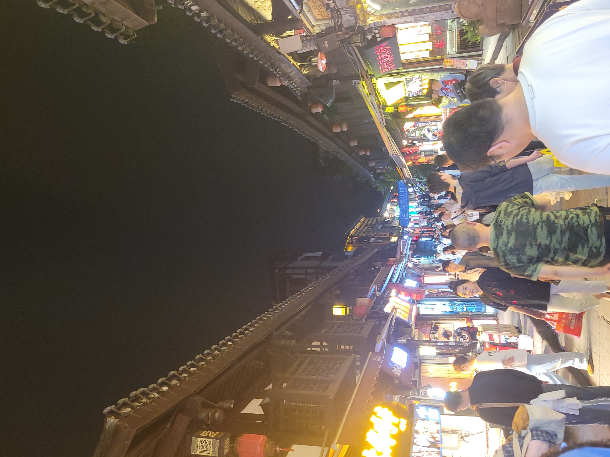 A nighttime street scene in Shapingba District, China, features a crowded pedestrian walkway lined with shops and restaurants illuminated by bright lights and signs in Chinese characters. A mix of people walk along the walkway, including individuals wearing casual clothing such as a white shirt, a camouflage shirt, and a black shirt. The architecture includes traditional Chinese-style buildings with ornate roofs, lanterns, and decorative elements.