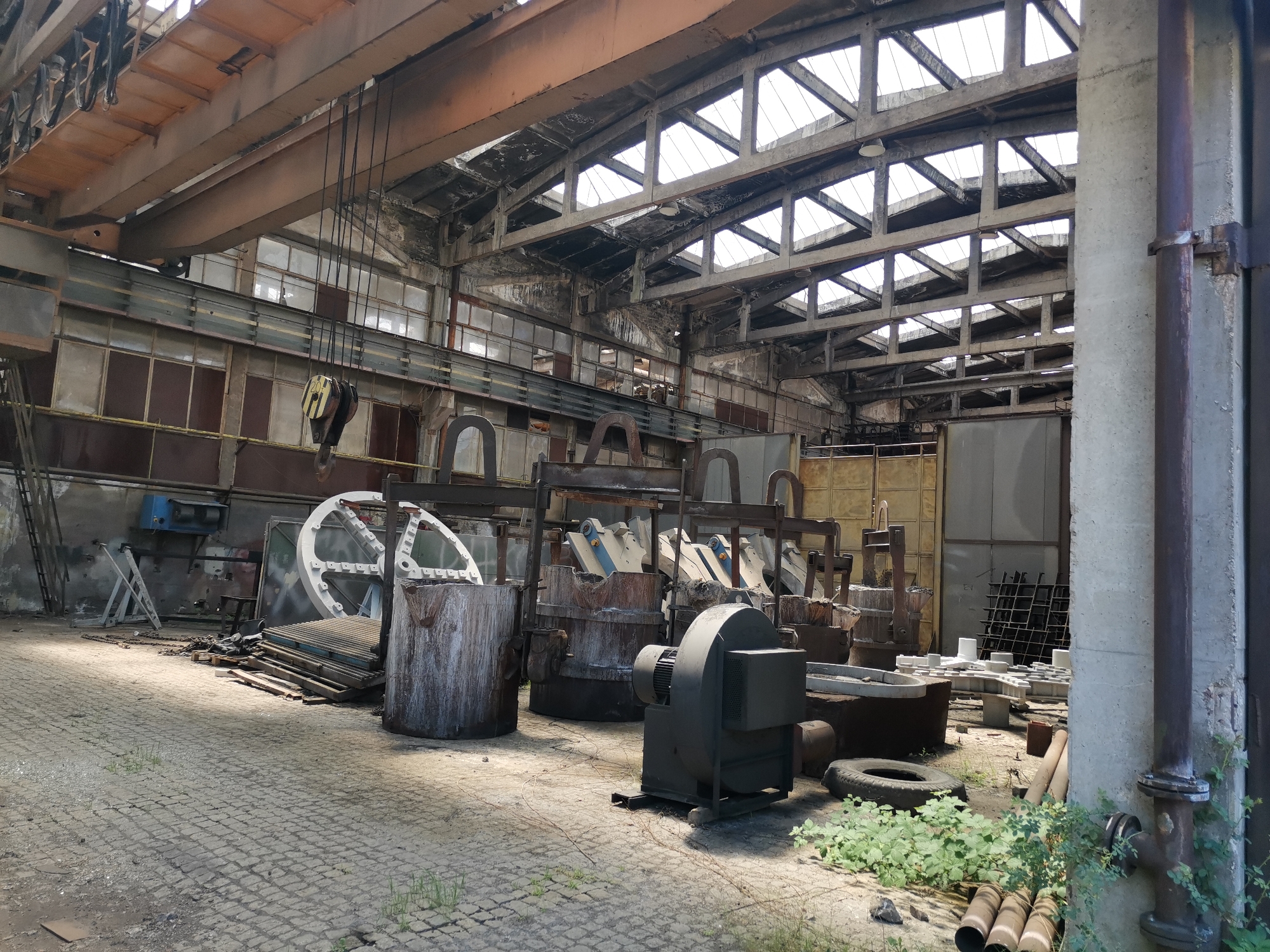 The image depicts the interior of a large, dilapidated industrial building in Verinsko, Bulgaria. The space is filled with heavy machinery and equipment, including large metal containers or crucibles, a blower, pipes, and a large white wheel-like structure. Sunlight streams through the gridded skylights in the roof, illuminating the concrete floor which is littered with debris and overgrown with vegetation in some areas. An overhead crane system is visible near the roof. The overall impression is one of disuse and abandonment.