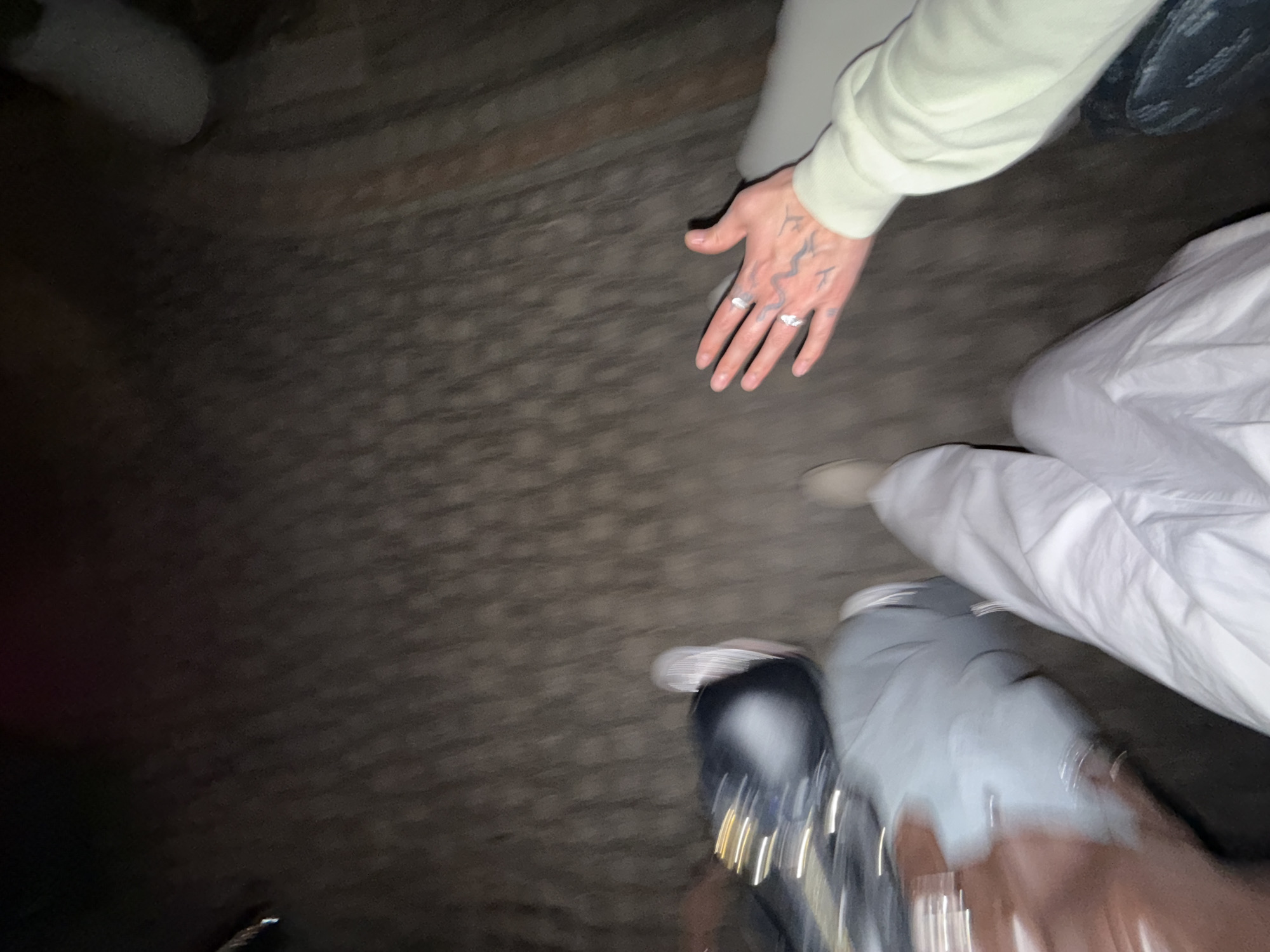 The image shows a low-angle view of the ground with several people standing in close proximity. One person's hand with tattoos and rings is prominent, positioned near the center. A person wears a light green long-sleeved shirt, while another is wearing white pants. The clothing of other individuals includes grey pants and a brown jacket. A dark bag or purse with silver embellishments is present in the lower right. The flooring appears to be asphalt or pavement. The location is unknown and there is no indication it is Yerevan, Armenia.