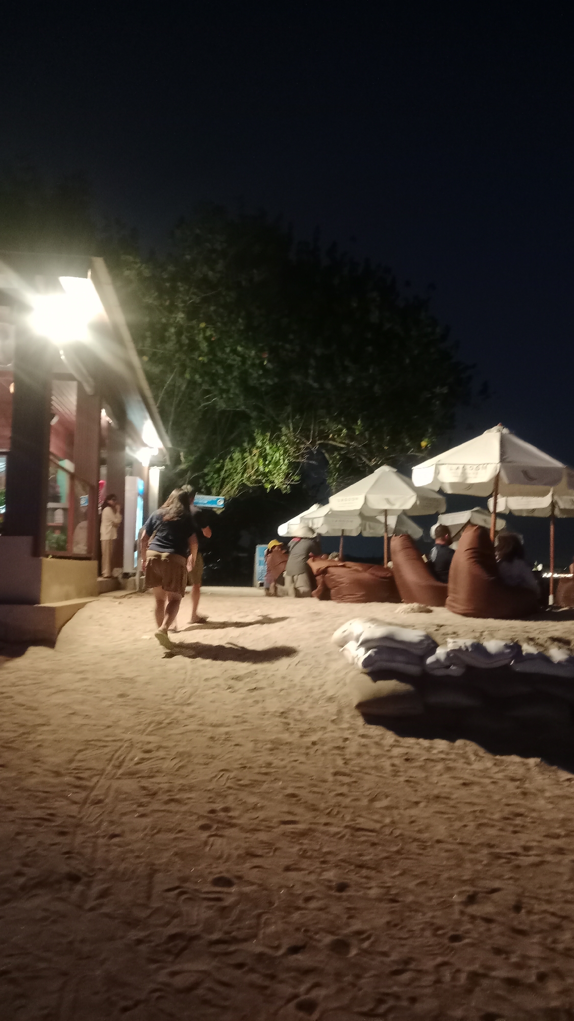 At night, a beach in Kuta, Indonesia, is illuminated by artificial lights. Several people are present. In the foreground, a woman with long dark hair is walking away from the camera on the sand. She is wearing a dark t-shirt and light brown shorts. Further back, there are beanbag chairs and umbrellas set up, with people sitting on the chairs. To the left, there is a building with lights on, and a person standing near the entrance. The sand is marked with footprints.