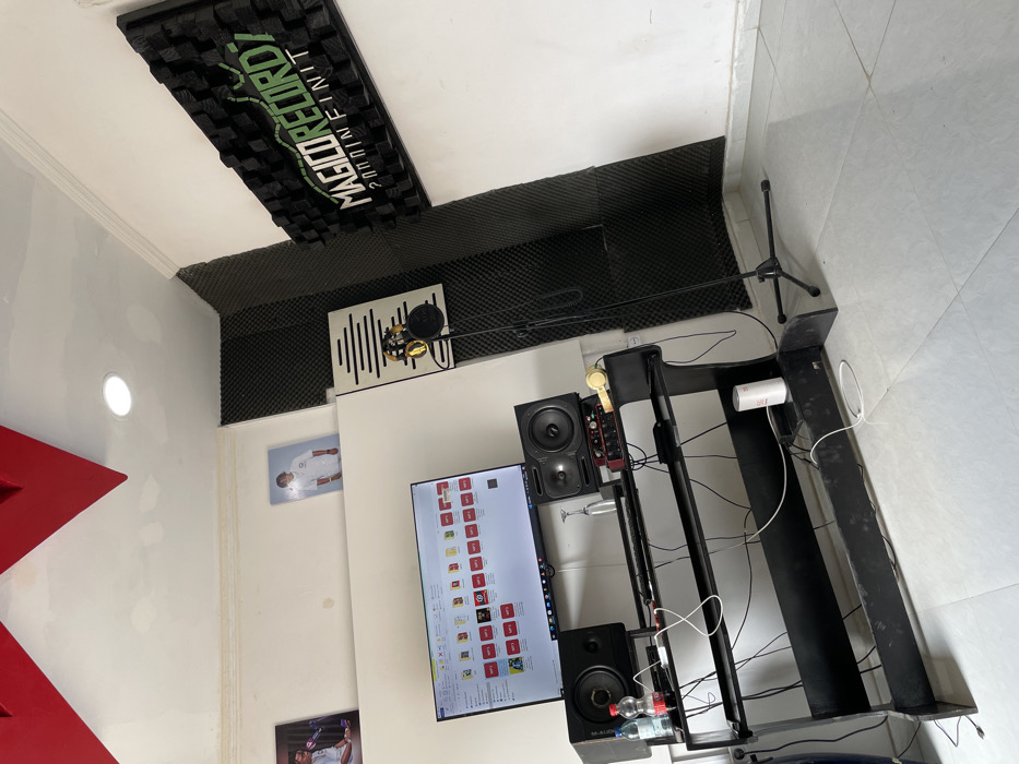 This image presents an indoor recording studio or music production setup. The room features white walls, with extensive black acoustic foam panels covering portions of the walls and along the wall-ceiling boundaries for sound treatment. A prominent, large rectangular acoustic panel with raised patterns and green text, "MAGO RECORDZ 200 INFINITE," is affixed to the ceiling.
At the center of the workspace, a professional microphone with a pop filter is mounted on an arm, positioned in front of a white acoustic panel featuring a vertical wave-like design. Below it, a black desk or workstation holds a large computer monitor displaying numerous icons, some labeled "SOLD," alongside two studio monitor speakers, one branded "M-AUDIO." A red "Focusrite" audio interface and other audio equipment are also arranged on the desk. Numerous cables are visible, connecting the various electronic components. On a lower shelf of the desk, two water bottles and a white cylindrical object are present.
To the right, a black microphone stand with a tripod base rests on the light-colored tiled floor. The white walls also feature decorative elements, including two framed pictures; the upper one shows a person in a white sports jersey. A bright circular recessed light is visible on the left wall, alongside a large red architectural element partially seen on the far left. The setting is equipped for audio recording and production, though no activity is currently in progress, and no individuals are visible. The lighting appears to be artificial.