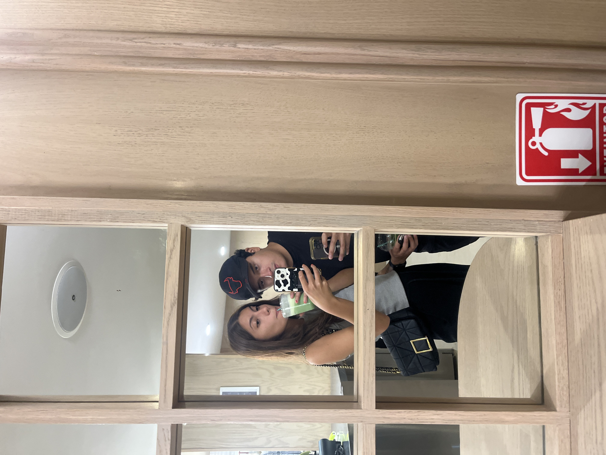 The image shows a reflection in a multi-paneled mirror. A couple is reflected in the mirror, appearing to take a selfie. The woman is drinking from a green beverage in a plastic cup. The man is wearing a black baseball cap with a red logo. They both are holding cellphones. A black quilted handbag with a gold clasp is visible on the woman's shoulder. The mirror is framed in light-colored wood. A fire extinguisher sign is affixed to the upper right corner of the frame. The interior decor suggests a commercial establishment, potentially in the city of Puebla, Mexico, although specific visual indicators of the city are not present.