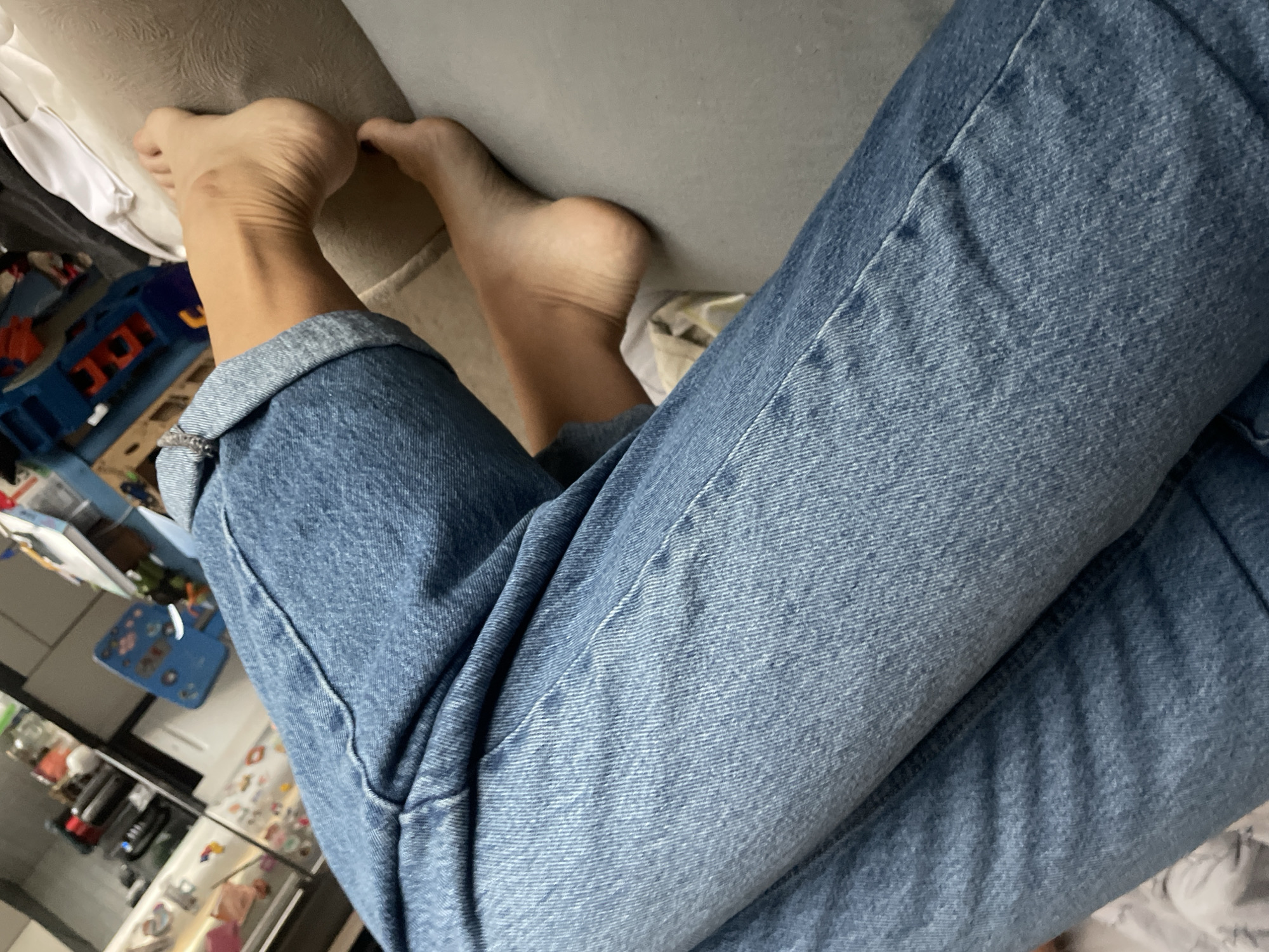 The image shows a person wearing blue jeans with bare feet resting on a light-colored surface, possibly a couch. The scene appears to be indoors, with a glimpse of a kitchen and what seems like children's toys in the background. The image doesn't provide any information to specifically confirm its location in Baranovka, Russia.