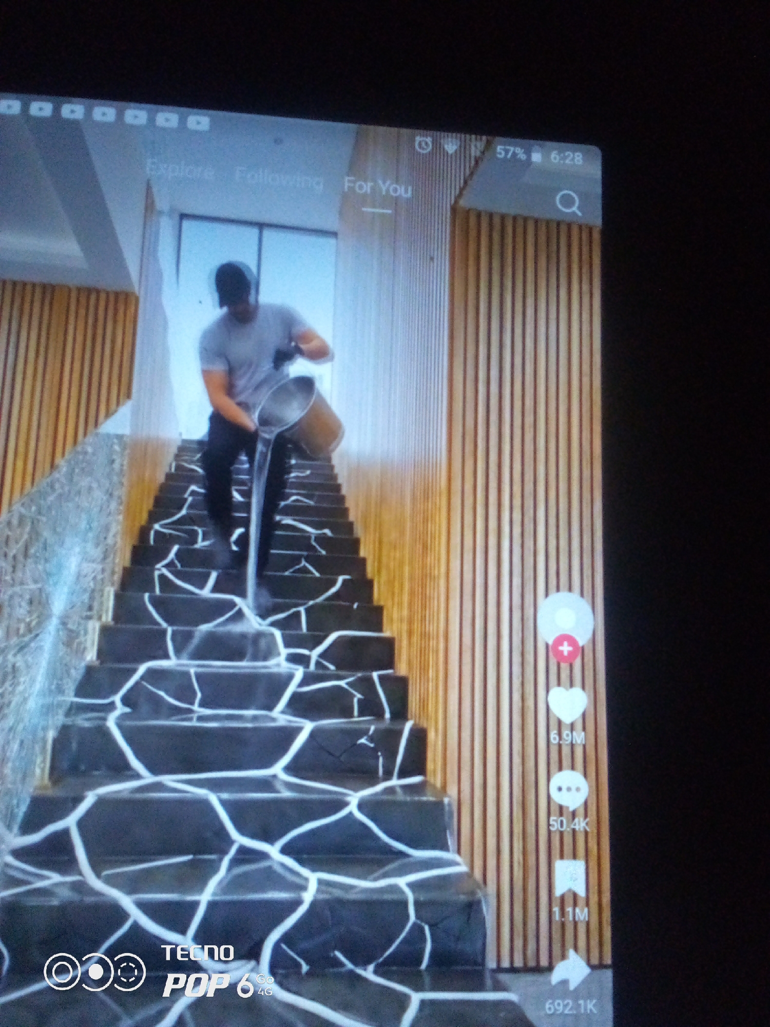 A person wearing a gray t-shirt and dark pants pours water from a bucket down a staircase. The staircase is dark with white geometric patterns. Vertical wooden planks line the wall to the right of the stairs. A phone screen is visible in the foreground with social media icons and text. The location is identified as Howayi, Nigeria.