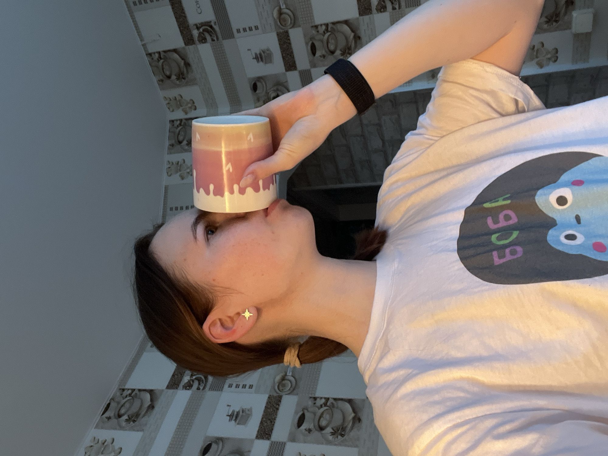 A woman is holding a mug to her forehead, seemingly balancing it. The mug is white with a light pink section and white drip-like patterns. The woman is wearing a white t-shirt with a graphic design on it. She has dark hair, pulled back into a low ponytail and fair skin. Her right ear is adorned with a gold star-shaped earring. She is wearing a black wristband. The background shows a wall covered in tiles with coffee-related patterns. The location is Chaikovsky, Russia.