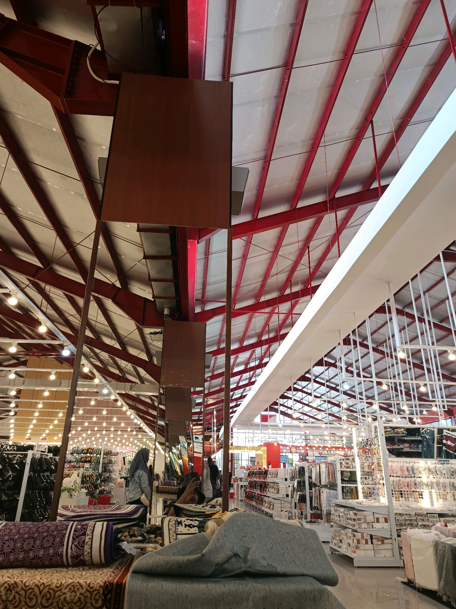This image depicts the interior of a large retail store in Bulukumba, Indonesia, likely a department store or a hypermarket, characterized by its expansive ceiling structure. The ceiling features exposed red metal beams forming a truss system, supporting a translucent white roof paneling that allows diffused light to enter. Numerous pendant lights hang from the ceiling, illuminating the store's interior.
In the foreground, several rolled carpets and fabric items are displayed on low platforms, suggesting a home goods section. Moving further into the store, rows of shelves are visible, stocked with various merchandise. To the left, displays of footwear and clothing are apparent. In the center and right of the image, more shelving units showcase a variety of products, including what appear to be accessories, textiles, and packaged goods.
Several individuals are present within the store. In the mid-ground, a woman wearing a gray hijab and a light-colored jacket stands near a display. To her right, another person, likely a man, is partially visible, holding a smartphone as if taking a photo or video. Further into the store, other shoppers and staff members are seen among the aisles. The overall environment suggests a well-lit, organized commercial space designed for high-volume retail.