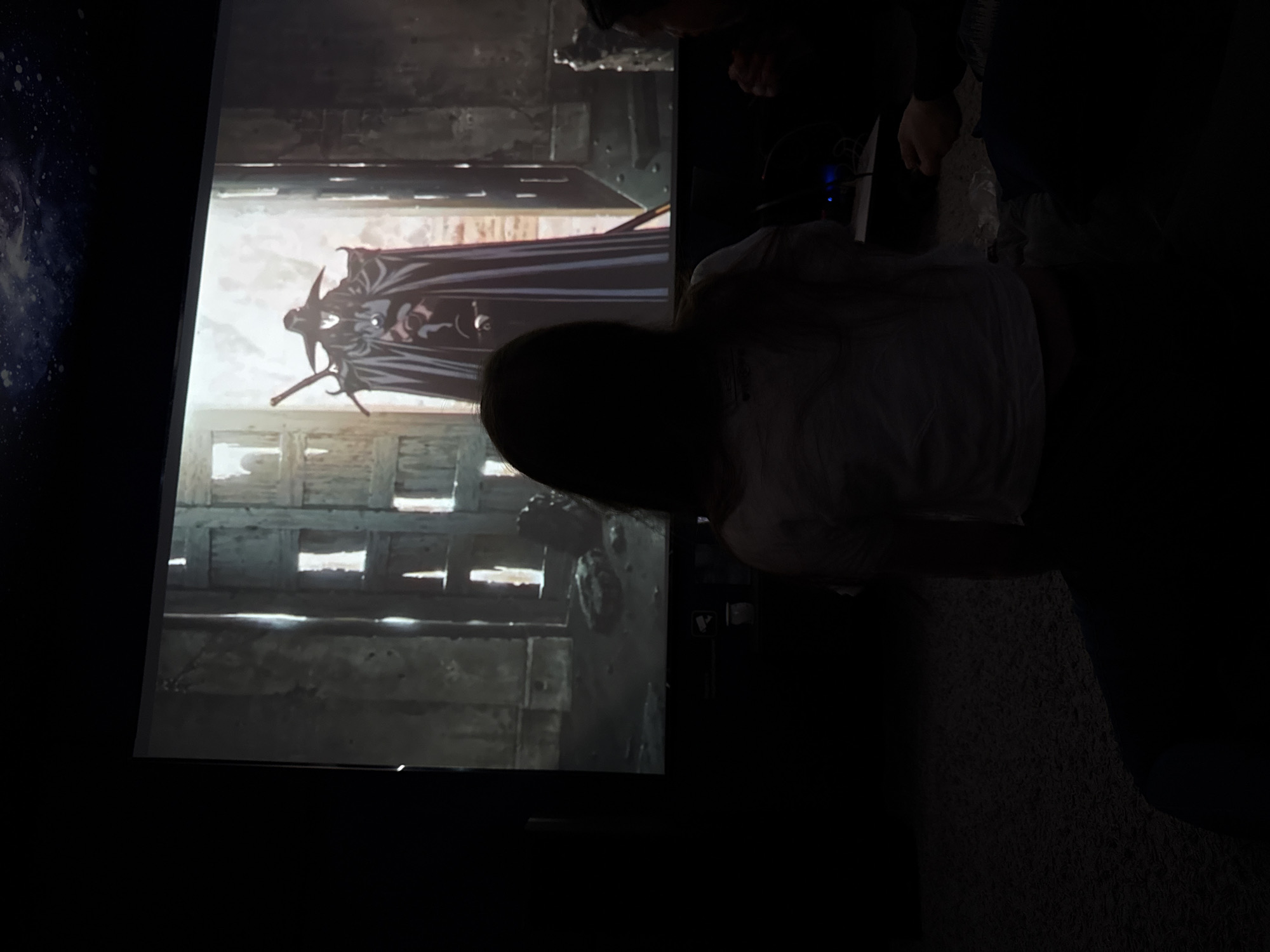 The image shows a screen displaying a dark fantasy scene with a figure in a long cloak holding a sword. The scene appears to be set in a dilapidated, possibly medieval, environment. Two individuals are partially visible in the foreground, obscuring part of the screen. The location is indiscernible.
