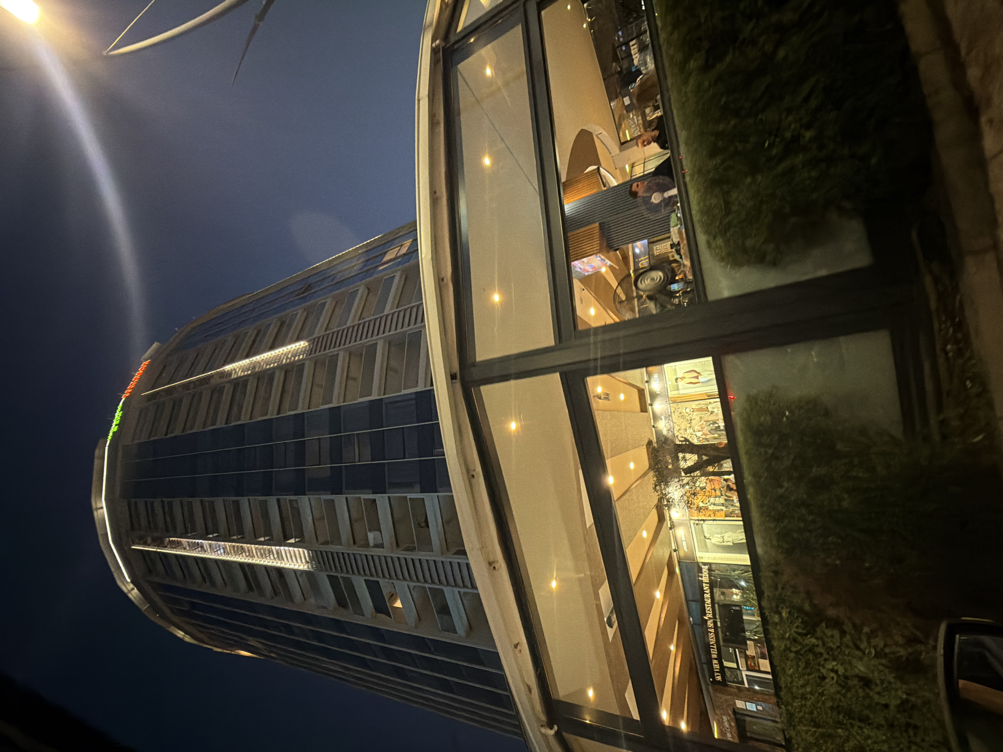A low-angle, wide shot captures a modern, curved, glass-fronted building at night, likely a restaurant or hotel, with a tall skyscraper dominating the background. The skyscraper, illuminated by vertical lines of light and a neon sign at its peak, has a repeating pattern of balconies and windows. Streetlights and the glow from within the glass-fronted building illuminate the scene. Inside the glass-fronted section, several people are visible, seated at tables and standing, with lighting fixtures and architectural details of the interior apparent. A hedge in the foreground partially obscures the lower part of the glass-fronted building. The sky above is dark and clear, with a bright light source to the upper right. The overall impression is one of urban nighttime activity.