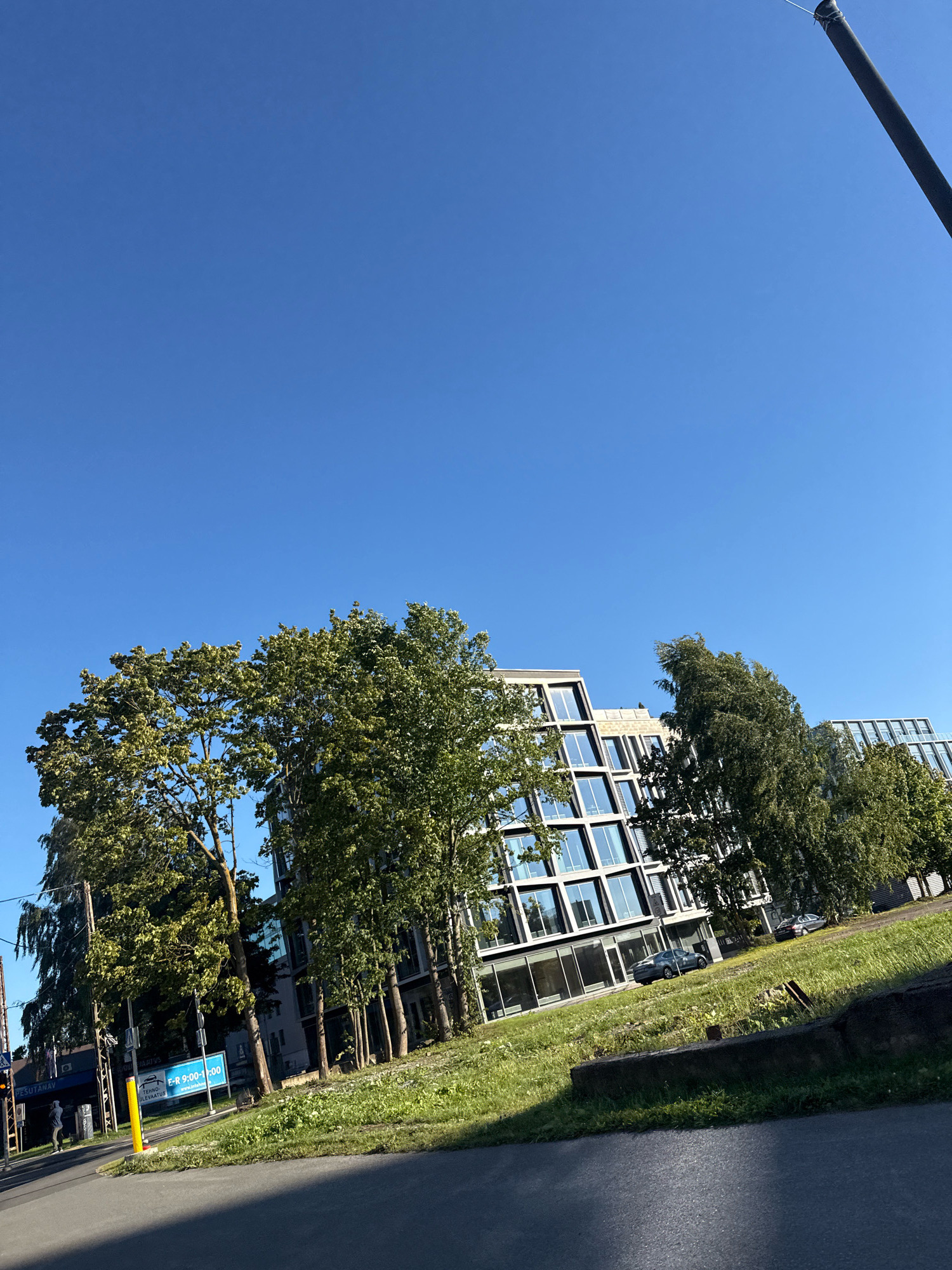 The image captures a sunny day in Tallinn, Estonia, with a clear blue sky. In the foreground, a paved surface with a shadow diagonally bisects the frame. Beyond this, a grassy incline leads up to a modern building complex. The building features a facade of glass windows and light-colored structural elements. To the left of the building, several mature trees with lush green foliage stand tall. A street is visible at the bottom left, with traffic lights, signage, and a few cars. One sign displays the text "E-R 9:00-18:00", indicating business hours. The scene suggests an urban environment, possibly an office or commercial district, bathed in natural sunlight.