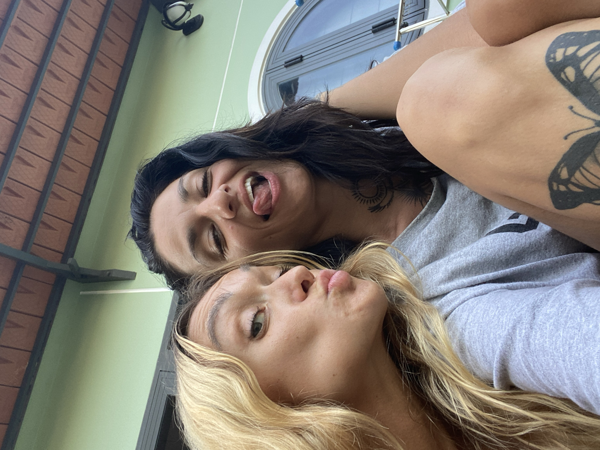 In Mũi Né, Vietnam, two women are captured in a selfie outdoors. The woman on the left, with blonde hair, is puckering her lips towards the camera. The woman on the right, with dark hair, is sticking out her tongue and smiling with her eyes closed. She has a tattoo of a sun-like symbol on her chest and a butterfly tattoo on her thigh. They are both wearing casual grey t-shirts. The background features a light green wall, a section of a building's roof with reddish-brown tiles, and a large, arched window.