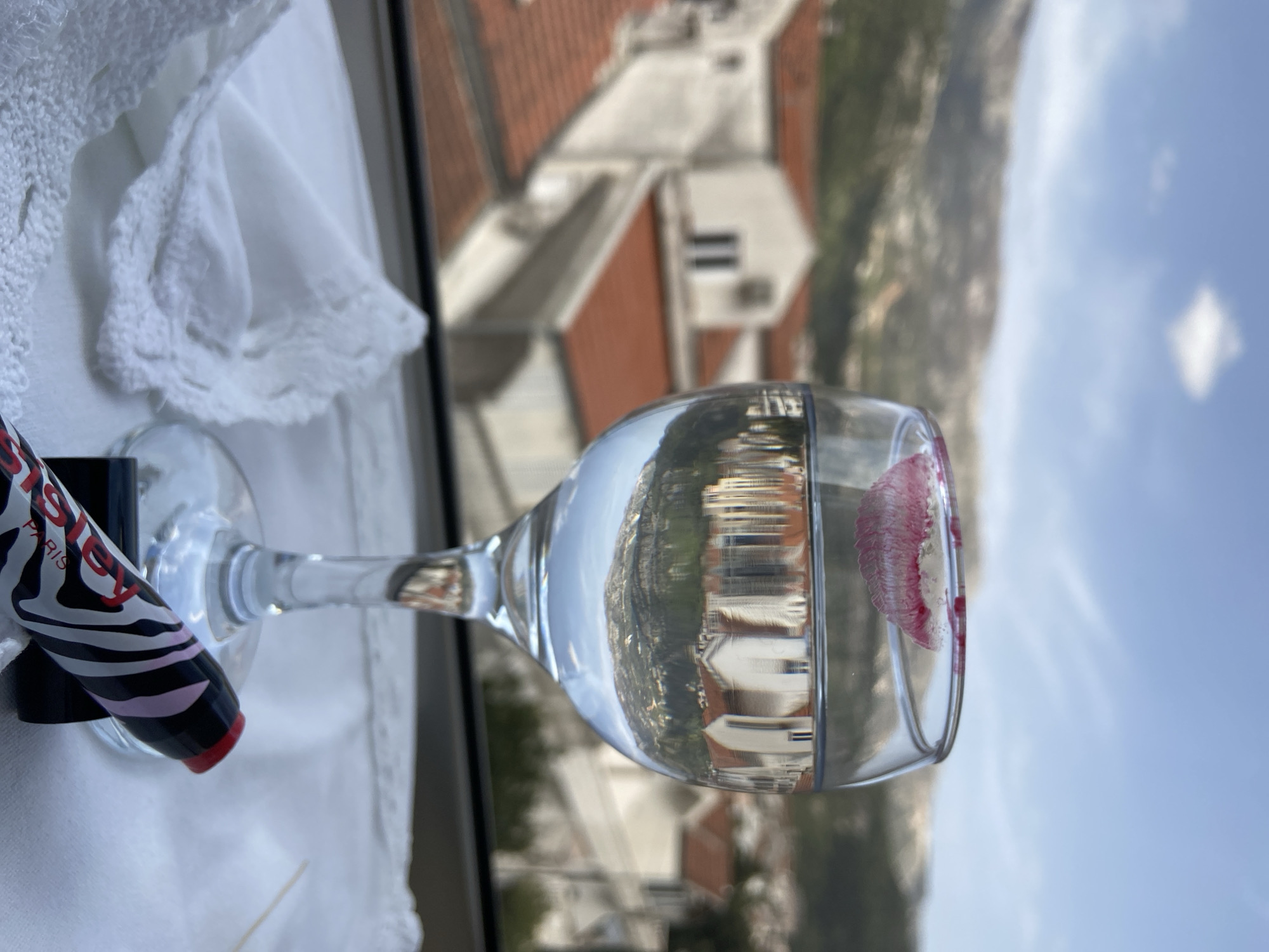 The image shows a wine glass placed near a window, likely inside a building. The glass contains water and a lipstick stain on the rim. A lipstick tube with black and white zebra print is lying beside the glass. The view through the glass and window reveals a cityscape with buildings, red-tiled roofs, and distant mountains against a blue sky with some clouds, consistent with Budva, Montenegro. A white, possibly lacy, cloth is situated next to the glass.