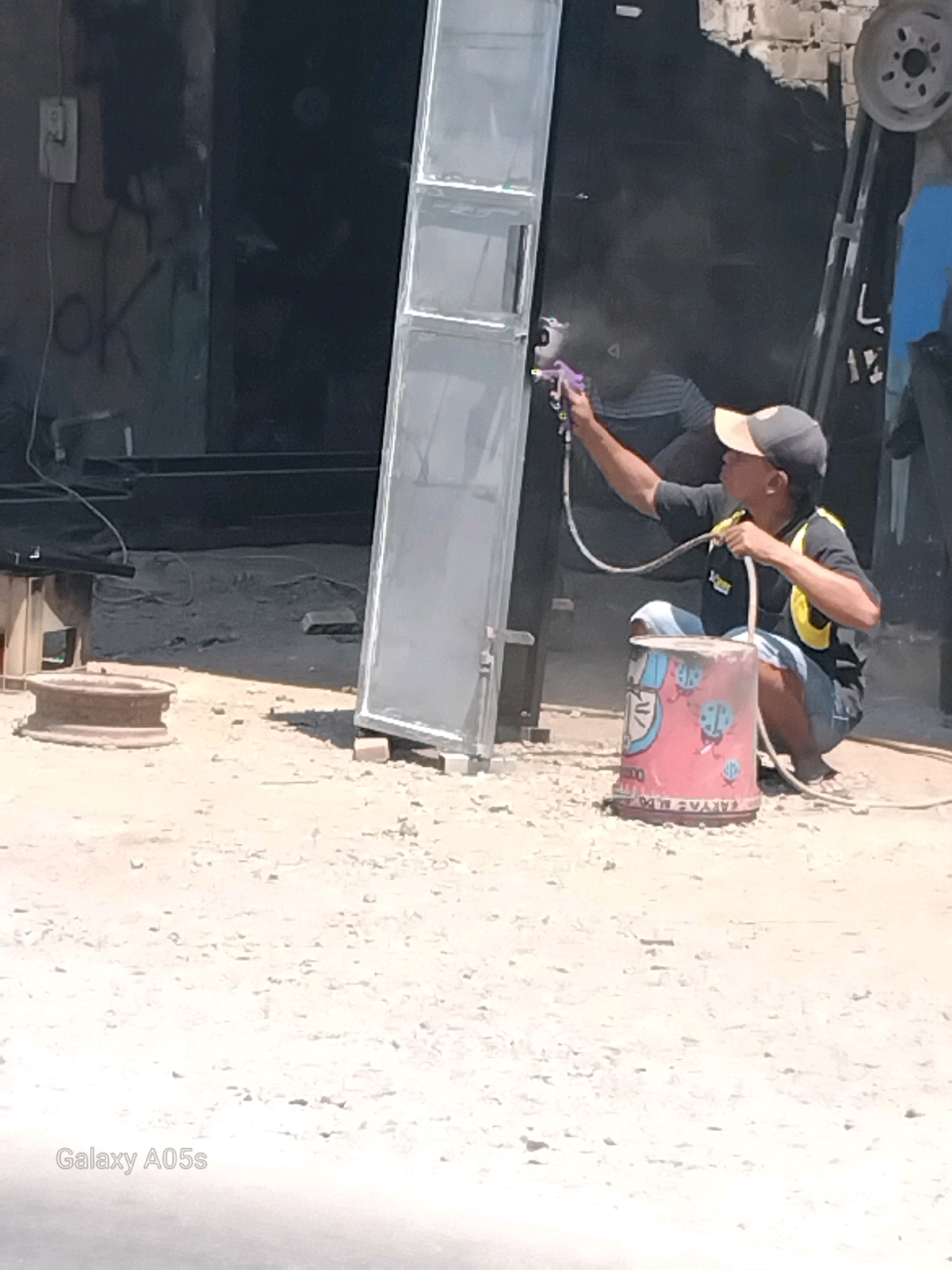 A person, wearing a baseball cap and a t-shirt with a yellow vest, is spray-painting a metal door. The person is crouching down next to a pink bucket with a cartoon design. The scene appears to be outdoors in what looks like a workshop or construction area. There is a large metal object, possibly another door or panel, leaning upright and being painted. The ground is covered in sand and small stones. In the background, there is a brick wall and some industrial equipment. The overall impression is of a DIY or small business operation. The watermark "Galaxy A05s" is visible in the lower-left corner of the image, suggesting it was taken with that particular phone model.