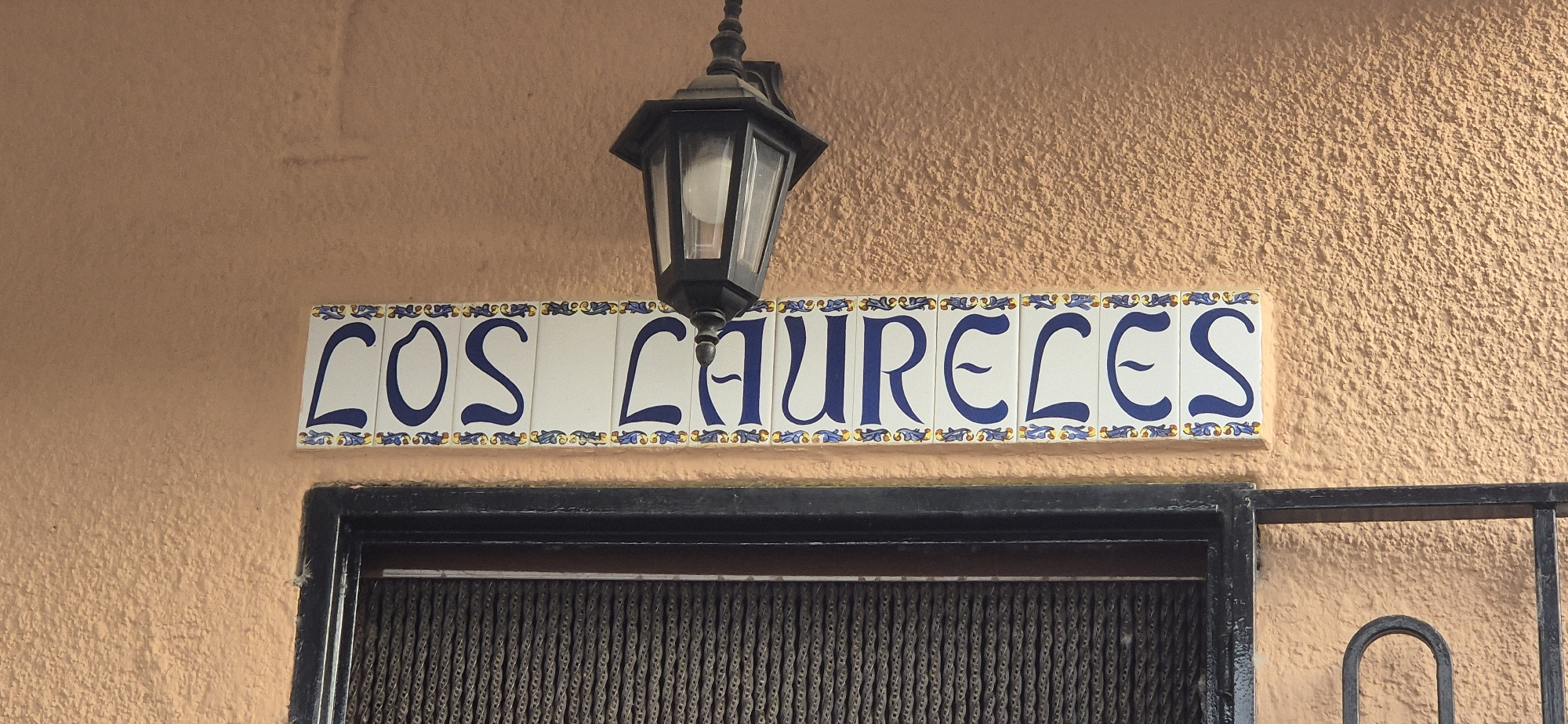 The image shows a close-up of a sign that reads "LOS LAURELES" in blue lettering on white ceramic tiles. The sign is mounted on an orange, textured stucco wall. Above the sign is a black, hexagonal lantern with glass panes. Below the sign is a dark-colored metal frame with a screen or gate behind it. The scene appears to be an exterior shot, possibly of a building entrance.
