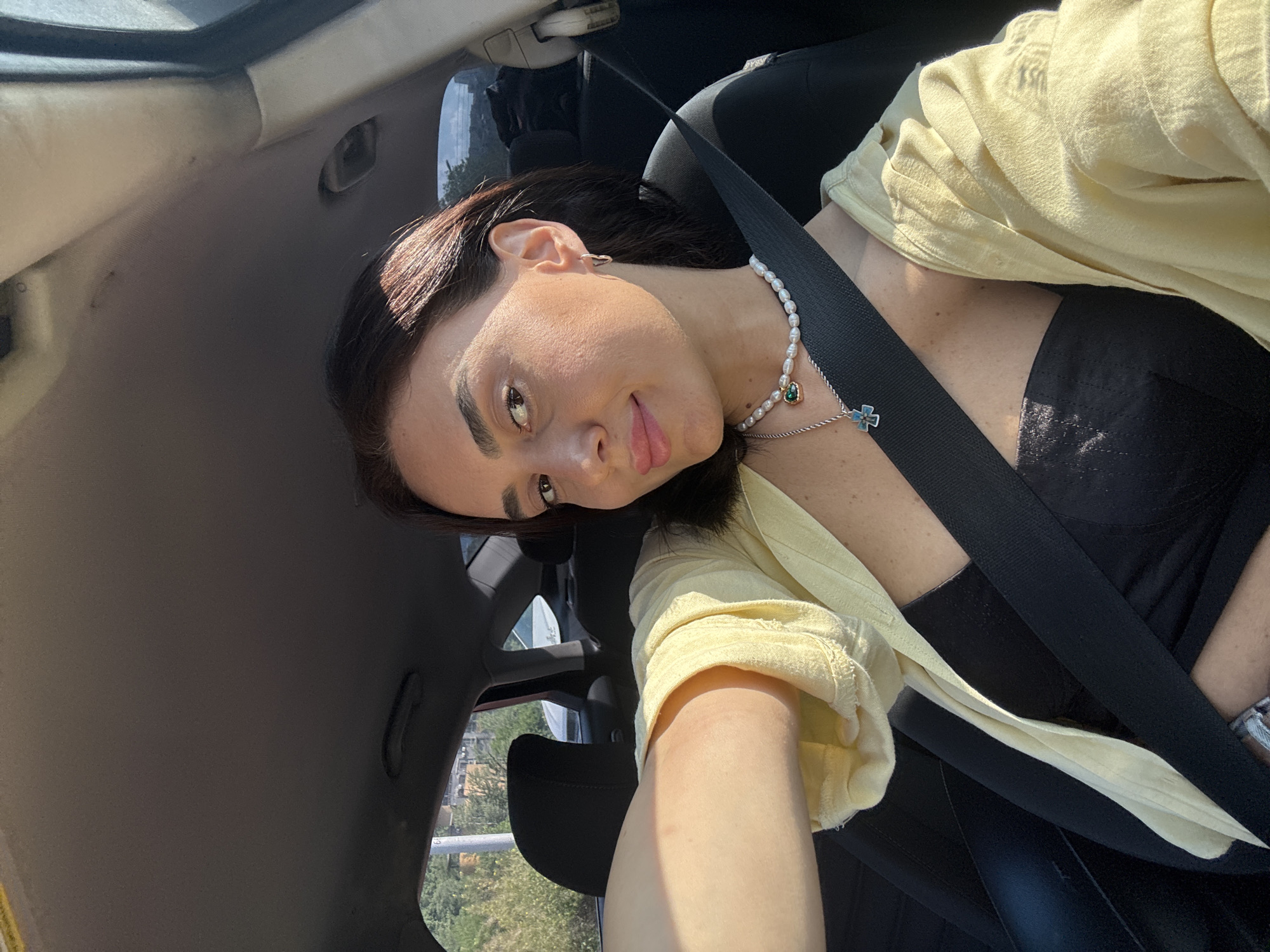 A woman with dark hair is seen from the chest up, inside a car, facing forward, with the driver's side window on her left. She is wearing a black strapless top, a light yellow open shirt, a pearl necklace with a green pendant, and a silver necklace with a blue cross. She has on a silver hoop earring in her left ear. The car's black seatbelt is diagonally across her chest. Sunlight streams in from the left, illuminating her face and arm. The background shows the interior of the car, including a black headrest and the car's ceiling. Outside the window, trees and buildings are visible, suggesting an urban or suburban environment. The context of Tbilisi, Georgia, is not discernible from the image alone.