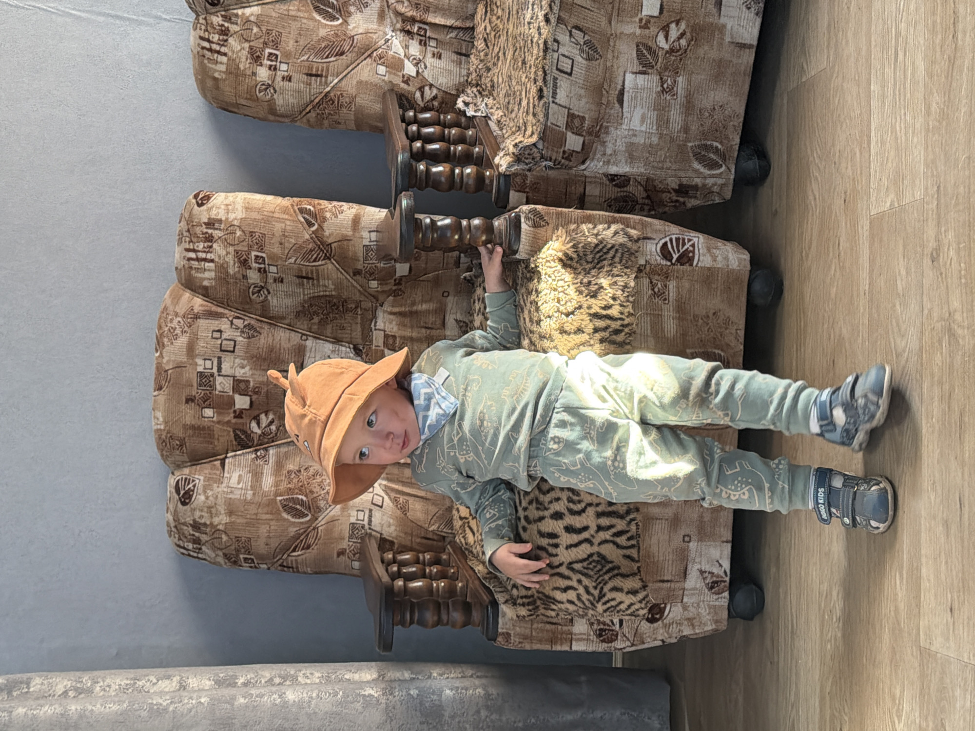 A young child, appearing to be a boy, stands on a wooden floor between two patterned armchairs. He is wearing a light green long-sleeved shirt and pants with a dinosaur pattern, an orange hat with a fin-like protrusion, a blue and white patterned bandana around his neck, and blue sandals. The armchairs are upholstered in a brown fabric with a repeating abstract pattern of leaves and squares. The child's left hand rests on the armrest of the armchair to his left. The wall behind the armchairs is painted a light gray. The image is taken indoors, likely in a residential setting in Красный Маяк, Russia, as suggested by the provided location information.
