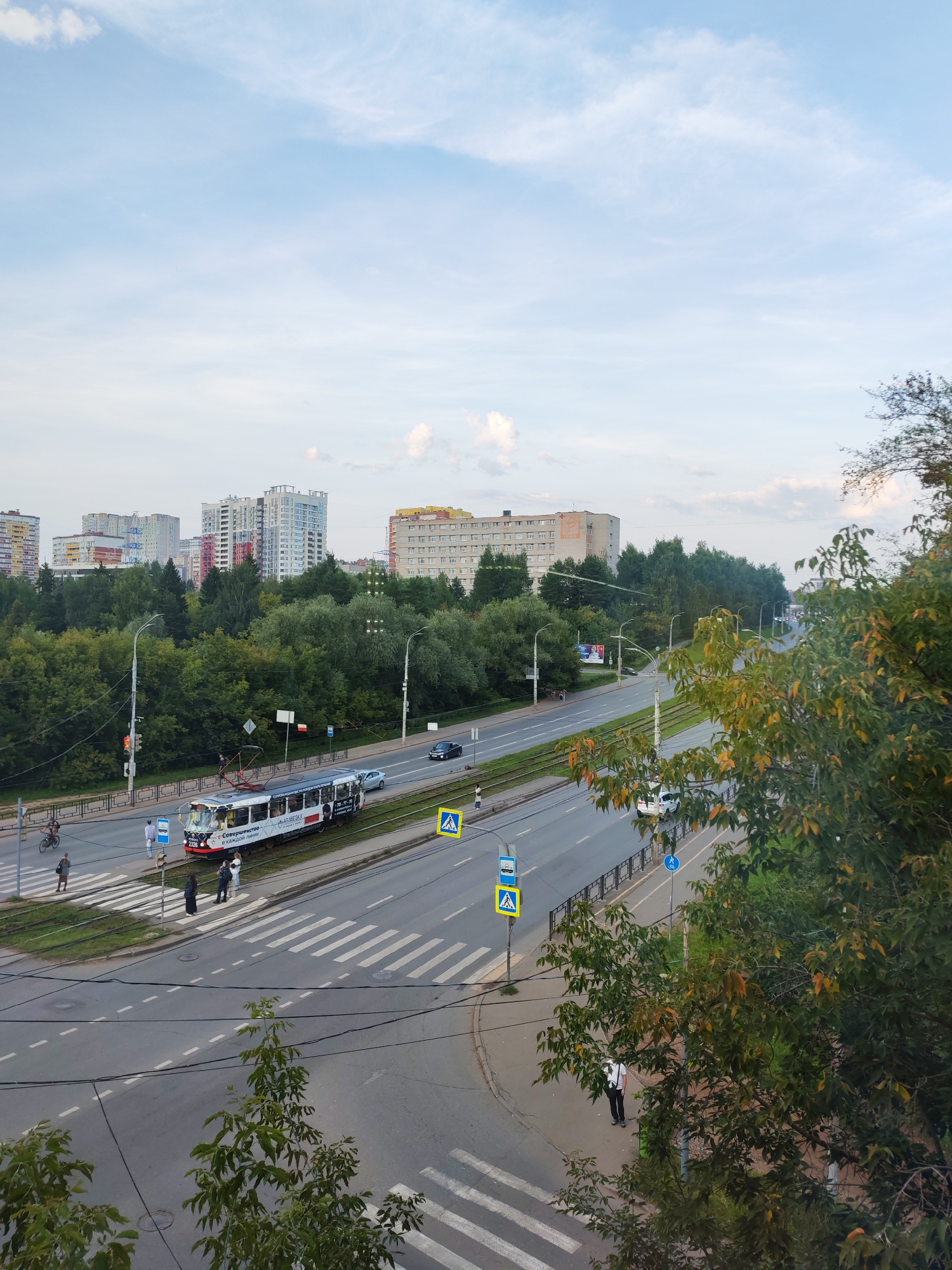 The image shows a view of a street in Izhevsk, Russia, with a tram moving along a track. The street is lined with trees and buildings, and there are pedestrians and vehicles present. The sky is partly cloudy. The tram appears to be modern and white with advertisements on its side. There are several crosswalks on the street, and traffic lights are visible. The buildings in the background vary in height and architectural style. Some trees in the foreground have leaves that are beginning to change color, suggesting it might be late summer or early autumn.