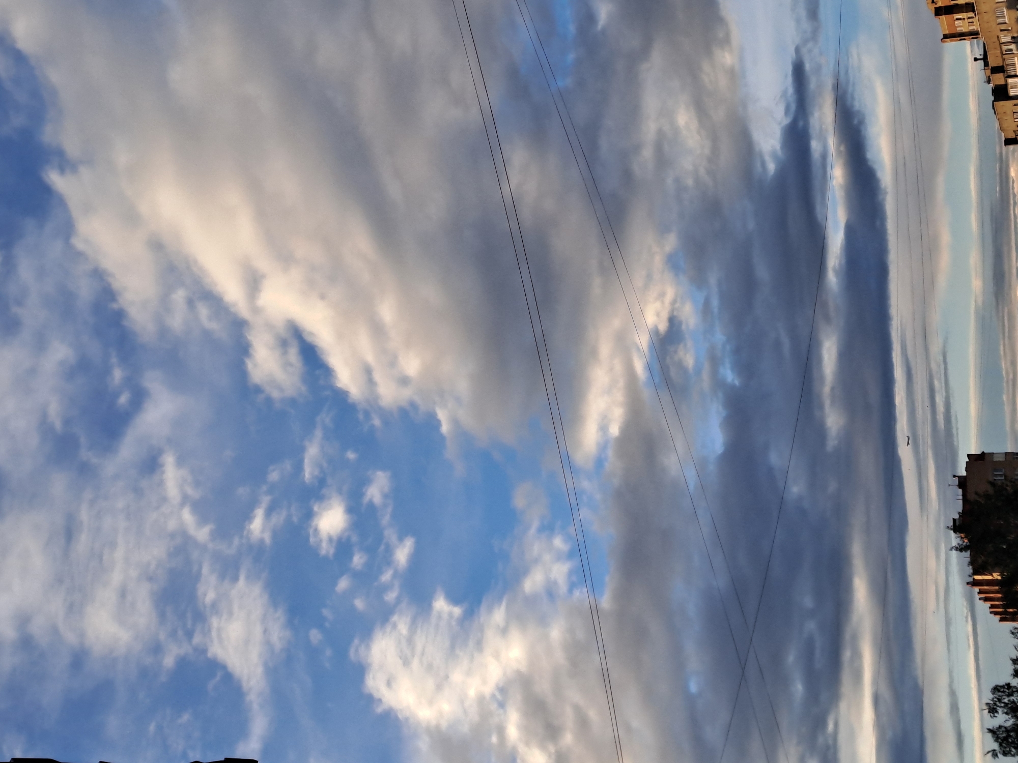 The image shows a sky filled with a mix of white and grey clouds with patches of blue sky visible. Three power lines diagonally bisect the frame. In the far right of the frame, the upper parts of buildings are visible, including what appears to be a residential apartment block. The buildings and some trees suggest an urban setting. The image was taken in Elektrostal, Russia. The angle suggests the camera was pointed upwards.